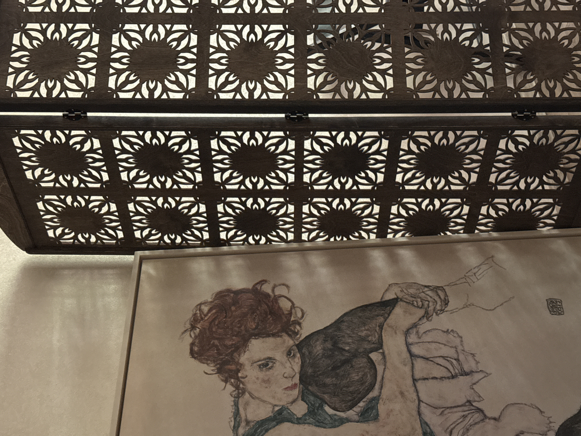 The image shows a close-up view of a painting hanging beneath a decorative, laser-cut wooden or metal structure. The structure features a repeating pattern of square panels with a central circular motif surrounded by stylized floral or leaf-like shapes. A hinge is visible on the structure. Below, a framed artwork depicts two people, possibly figures in an embrace. The visible figure in the artwork has red hair and is looking towards the viewer. The painting style appears to be expressive. The background behind the painting is a neutral color wall. There is no indication of the location being in Moscow, Russia.