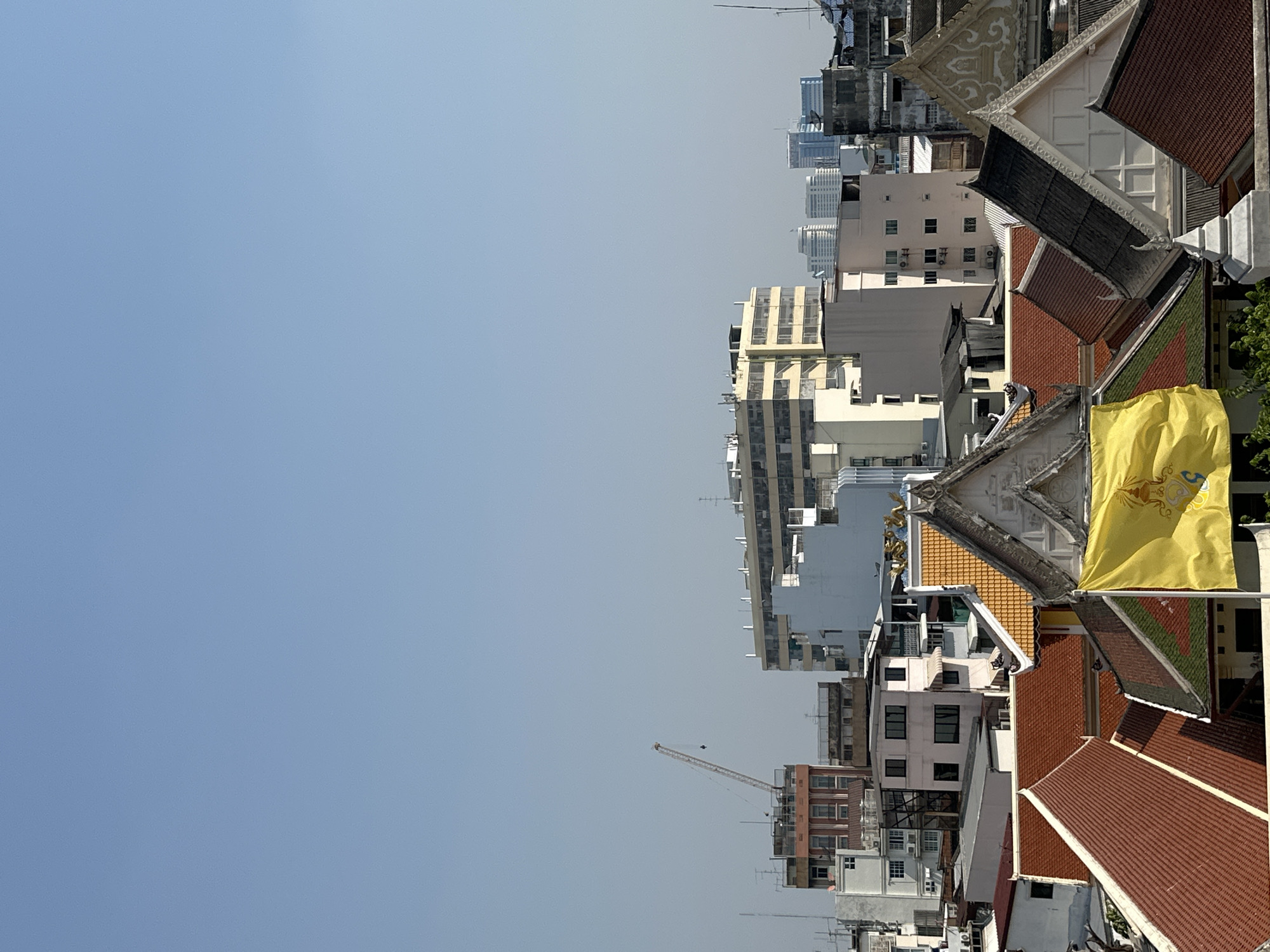 This image shows an elevated view of Bangkok, Thailand. The foreground features traditional Thai temple roofs with intricate designs, including a yellow flag possibly depicting the Thai royal family. In the midground, modern city buildings of varying heights are visible. A construction crane is present in the distance. The sky is a clear blue. The architecture juxtaposes the old and new of Bangkok.