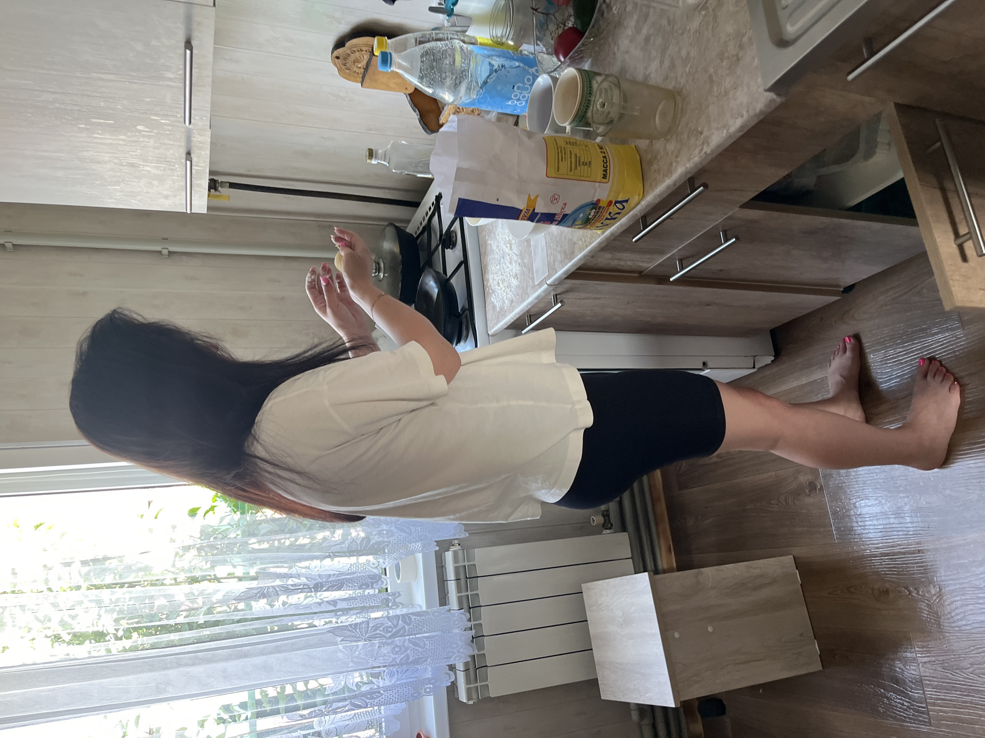 A woman with dark hair, wearing a white t-shirt and black shorts, stands in a kitchen, preparing food. She is holding small, round objects in her hands. The kitchen has light wood cabinets, a countertop with various items including a bag of flour and bottles of water, and a gas stove. A window with sheer curtains is visible in the background, allowing natural light to illuminate the scene. A white radiator is located beneath the window, and a small wooden stool is placed next to it. The floor is made of dark wood planks. The overall impression is of a domestic setting, possibly a home kitchen in Navahrudak, Belarus, given the context.