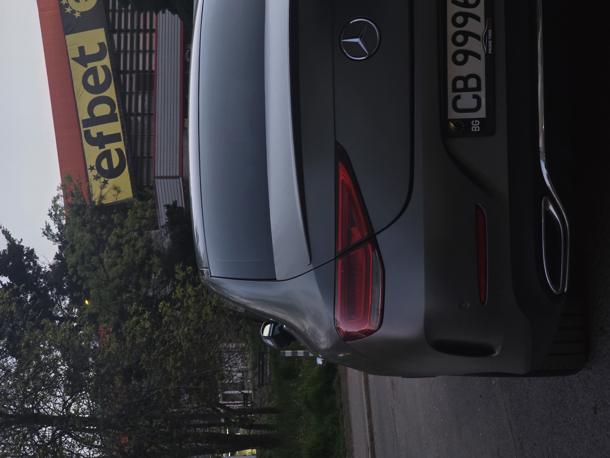 The image displays the rear-left quarter view of a dark gray Mercedes-Benz car, parked outdoors. The car features a prominent Mercedes-Benz logo on the trunk lid, sleek red LED taillights, and dual exhaust outlets integrated into the rear bumper. A Bulgarian license plate is clearly visible, showing the registration "CB 9996" with "BG" on the blue EU strip. Below the main numbers, the text "PRIME TIME AUTO" is discernible on the license plate holder.
In the background, to the left of the car, a large yellow and black banner with the word "efbet" and several stars is mounted on a building. The building appears to have a red facade and a structured roof. Numerous green trees and lush foliage are scattered in the background, both near the building and extending towards the right side of the frame. The car is situated on a paved surface, likely a road or parking area. The lighting suggests either dusk or an overcast day, with a bright sky providing soft illumination. The "efbet" banner and the Bulgarian license plate confirm the location as Sofia, Bulgaria. The image itself is rotated 90 degrees counter-clockwise.