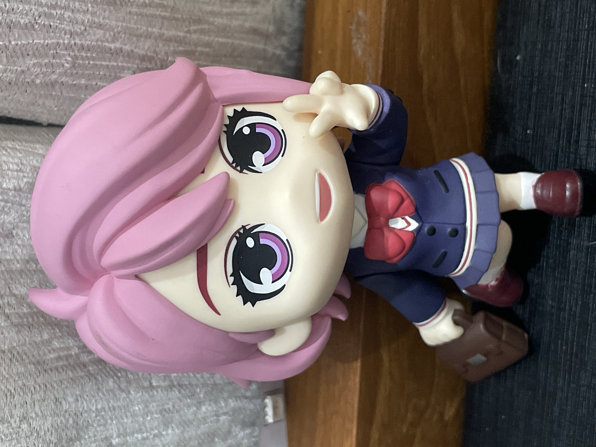 A close-up shot depicts a figurine of a female anime-style character with pink hair and large eyes. The figurine wears a school uniform with a red bow and holds a brown satchel. The figurine is situated on a surface with a gray textured background and a wooden element. The image was likely taken in an indoor setting. The location in the city of Denpasar, Indonesia, is not discernible from the image.