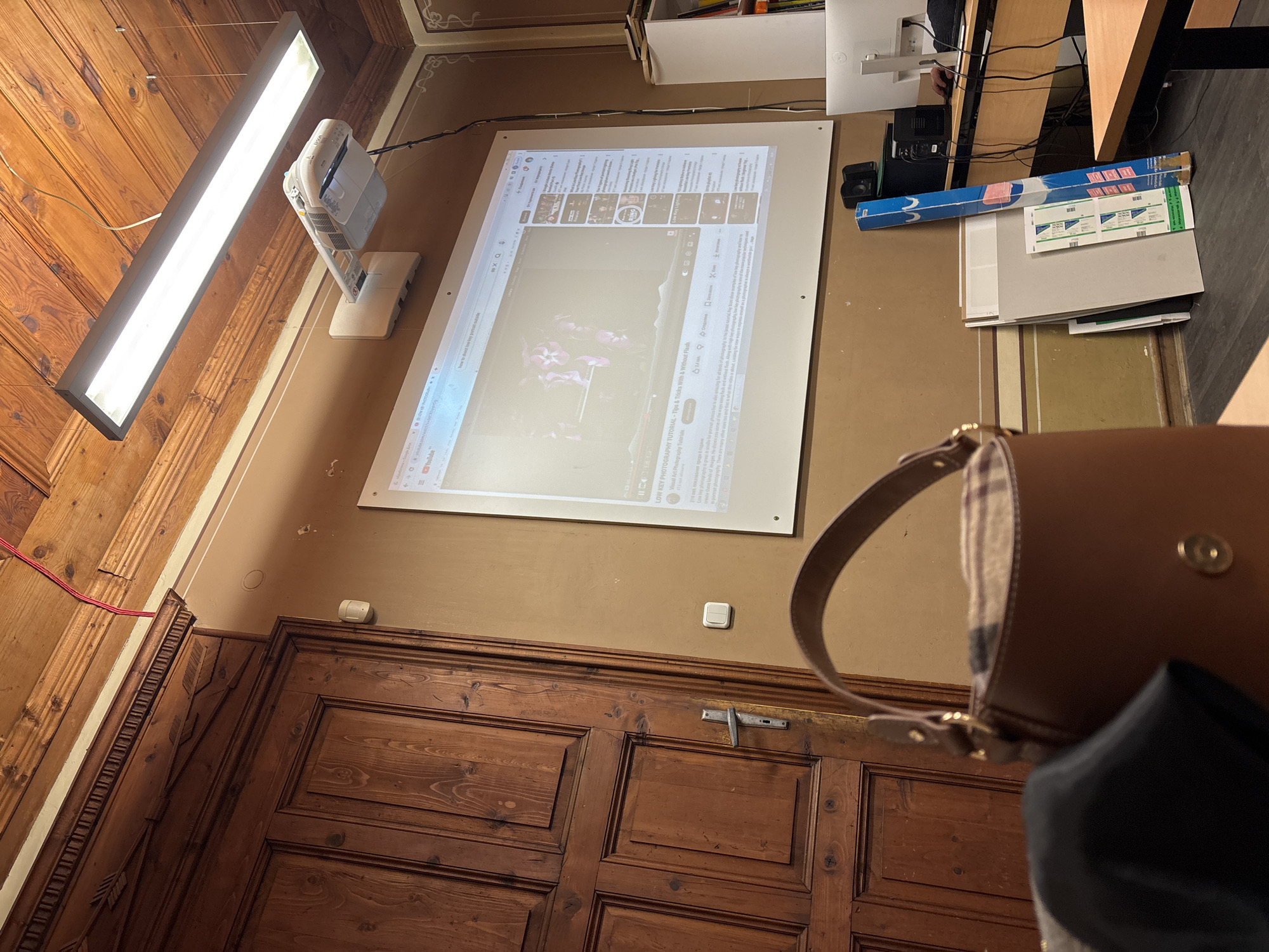 Here is a matter-of-fact description of the image:
The image shows an indoor scene, seemingly a room with wood paneling on the walls and a large wooden door at the bottom. A rectangular light fixture is mounted on the wall towards the upper left. Above the door and slightly to the right, a projector displays a YouTube video on what appears to be a screen mounted on the wall. The video seems to be about photography, specifically “LOW KEY PHOTOGRAPHY TUTORIAL.”
Various objects are visible on a table or shelf in the upper right, including what appears to be a computer monitor, boxes, papers, and other miscellaneous items. In the foreground, there is a partially visible brown purse or bag with a dark garment or cloth draped over it. The scene has a warm, yellowish tone.
