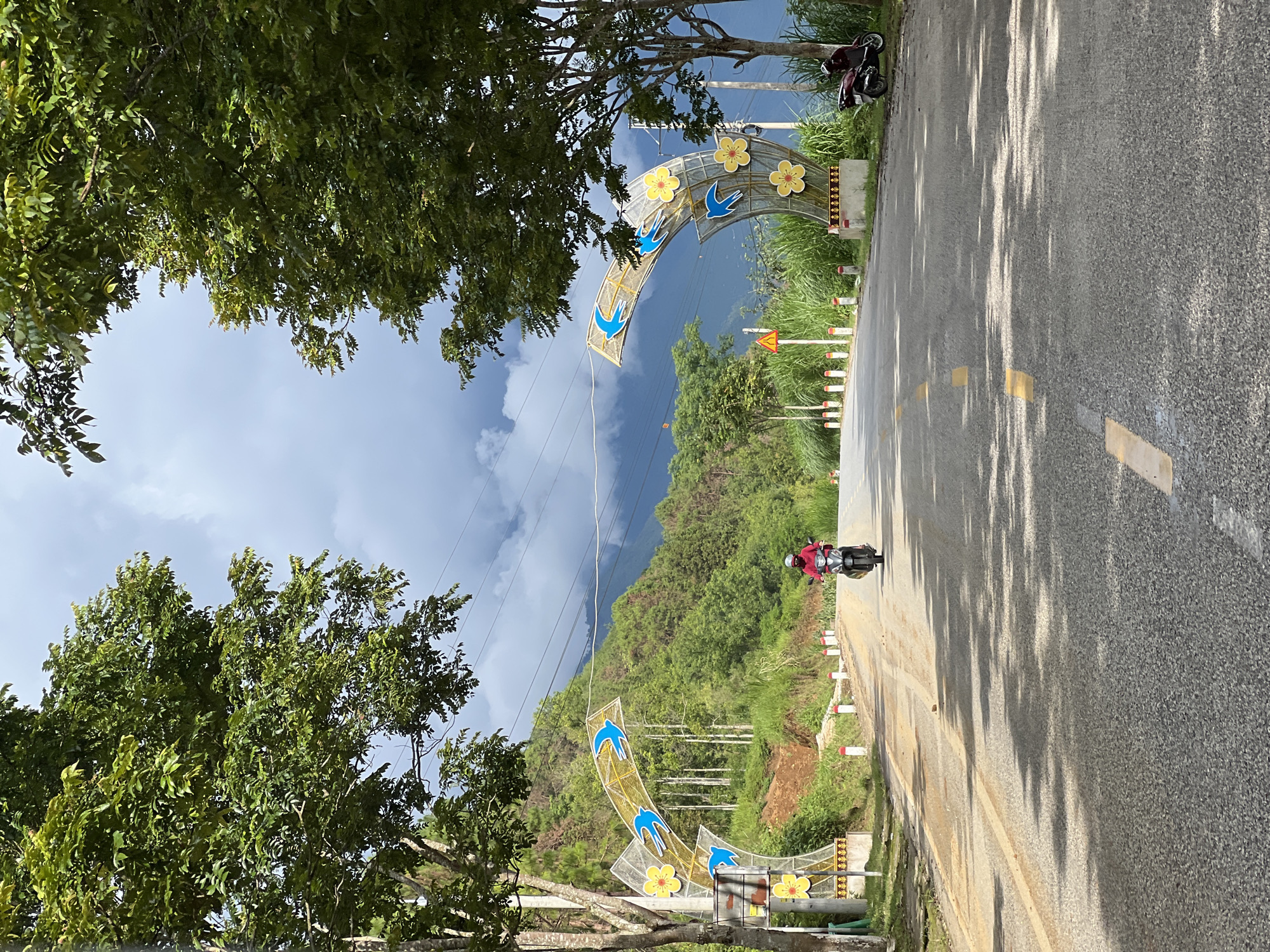 A wide-angle, eye-level shot captures a paved road in Quản Bạ Commune, Vietnam, framed by lush green trees on either side. The road slopes gently upwards and narrows into the distance, with yellow dashed lines marking the lanes. On the left and right sides of the road, decorative arches with yellow flowers and blue bird motifs are installed. A person on a scooter, wearing a red shirt and helmet, is traveling away from the camera on the right side of the road. In the background, a verdant, mountainous landscape is visible under a cloudy sky. A parked scooter is situated on the right side of the road near the decorative arch.