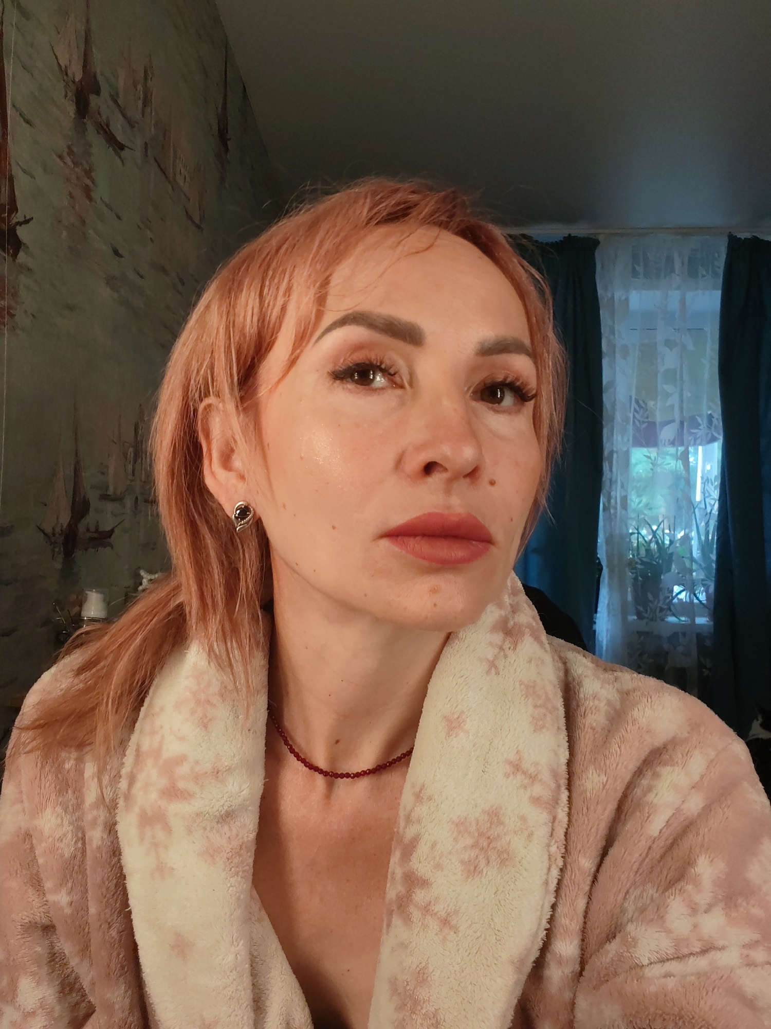 A woman with peach-colored hair and light makeup is posing for a selfie. She is wearing a fluffy pink and white patterned robe and a delicate beaded necklace. The background includes a wall with wallpaper featuring a nautical scene and a window with teal curtains. The scene appears to be an indoor setting, possibly a home, in Chaikovsky, Russia.