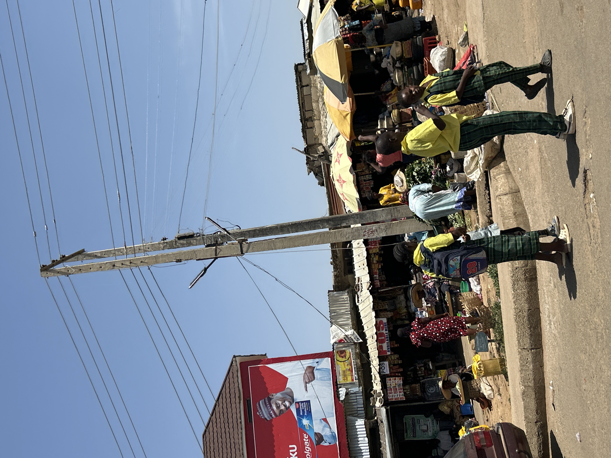 This image depicts a street scene in Jos, Nigeria. A concrete utility pole with numerous overhead wires dominates the left side of the frame. Adjacent to the pole is a building with a billboard advertising Colgate toothpaste, featuring a man in traditional attire. The right side of the image shows a busy market scene with vendors and customers under awnings and umbrellas. Two children wearing school uniforms (yellow shirts and green plaid pants) are walking along the sidewalk. A variety of goods are displayed for sale, including household items and foodstuffs. Several other people are present, including women in patterned dresses and straw hats. A car is visible at the bottom right of the frame. The sky is clear and blue.