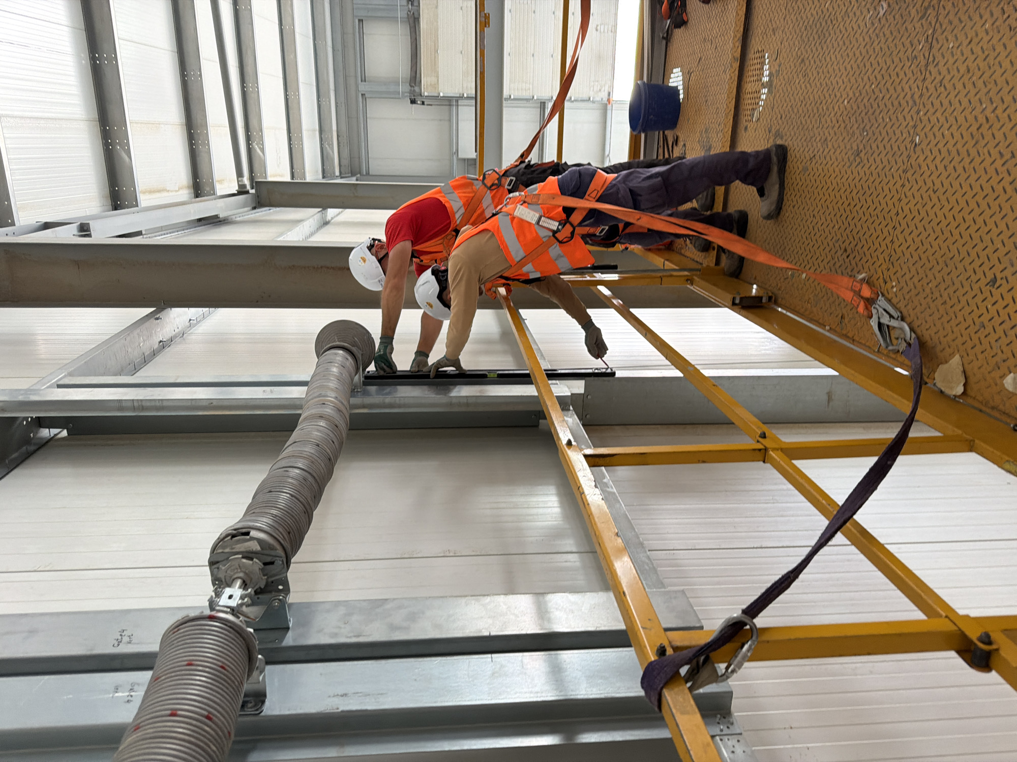 Two individuals wearing hard hats and safety vests are working on a large, coiled spring mechanism attached to a wall. One individual, in a red shirt and orange vest, is standing on a yellow platform and appears to be operating a tool. The other individual, in a tan shirt and orange vest, is also on the platform and is holding a pencil, possibly marking a measurement. The environment appears to be an industrial or construction site, with metal beams, corrugated metal walls, and a white ceiling visible. A blue bucket and tools are on the platform. The image is taken from a medium distance, capturing the upper bodies of the workers and the coiled spring.