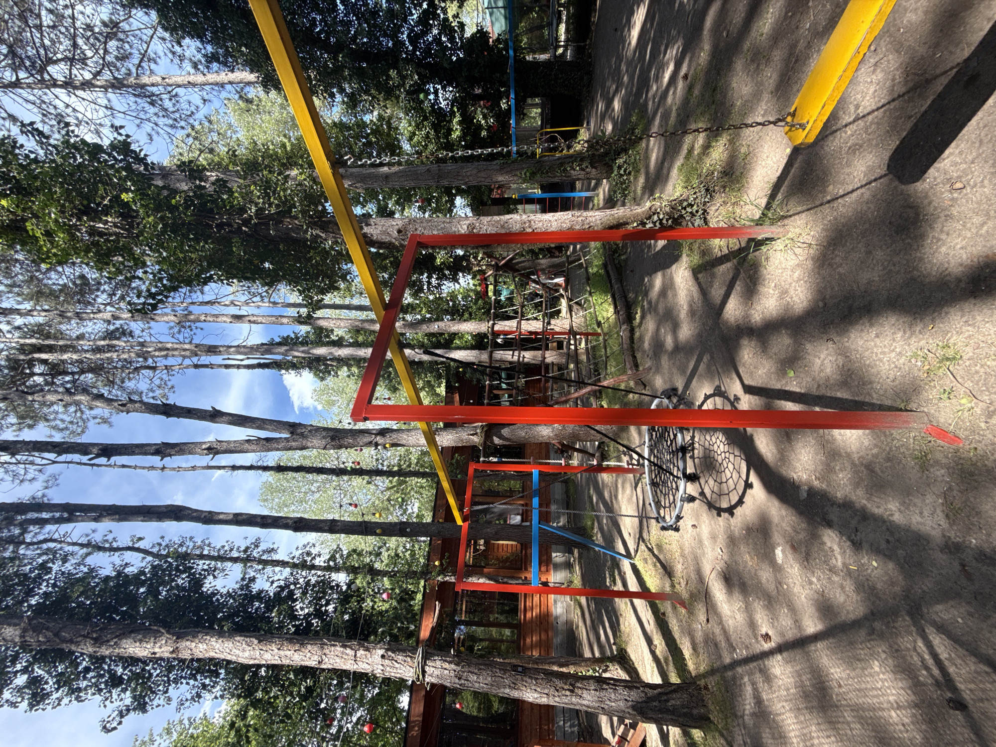 The image captures a children's playground located in the city of Novo Oriahovo, Bulgaria. The playground features wooden structures and rope bridges connecting various play areas among the trees. The equipment is painted in red, yellow and blue. The ground is concrete. The playground appears to be situated in a wooded area as mature trees provide a backdrop to the scene. There are no people visible in the image. The scene appears to be outdoors with bright sunlight.