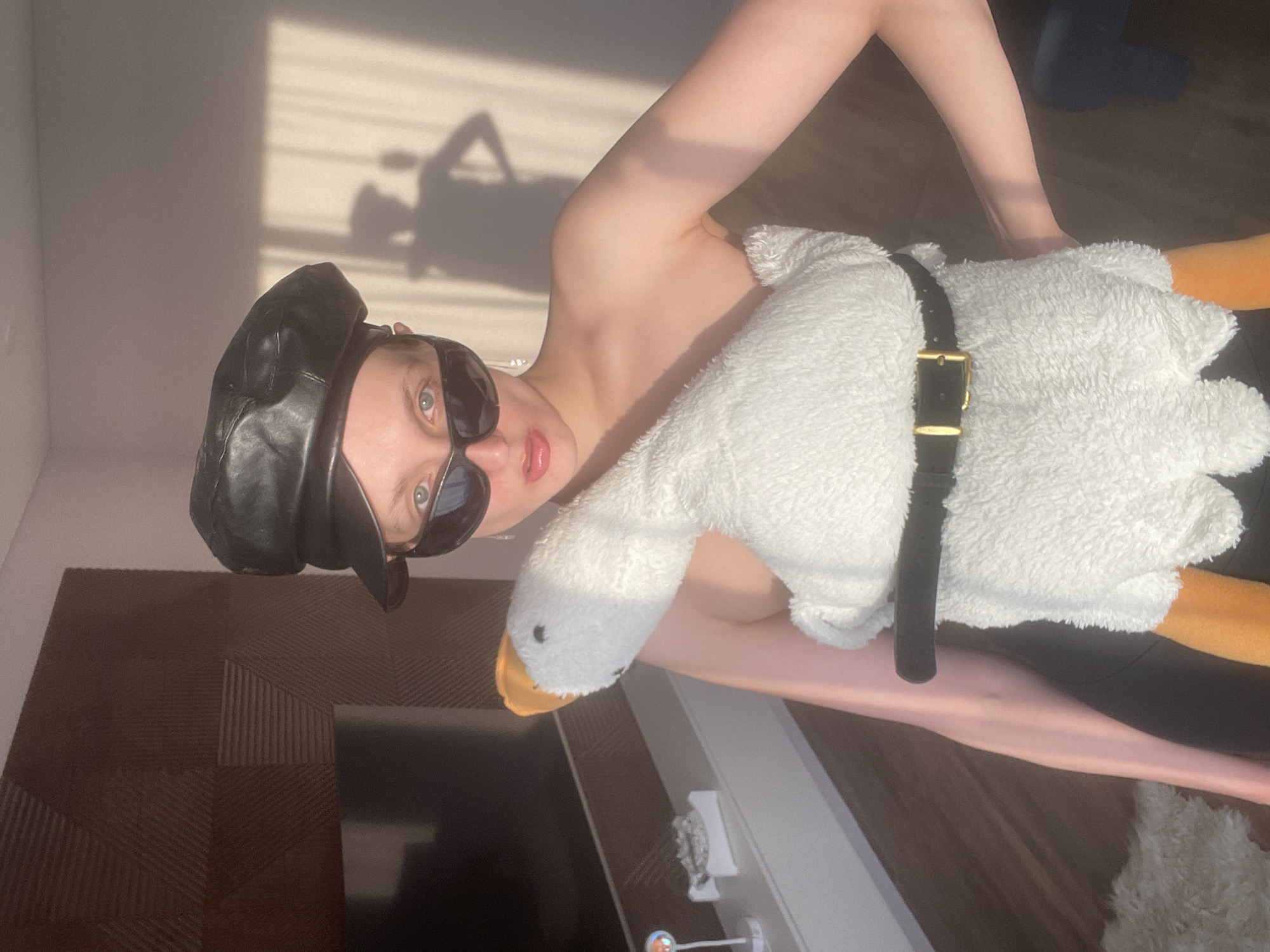 An indoor portrait captures a young person in Podshivalovo, Russia, posing playfully with a large plush goose toy. The individual, with light skin, wears dark sunglasses and a black, possibly leather, beret or flat cap. Their left shoulder and arm are bare.
Dominating the center of the frame is a large, white, fluffy goose or duck plush toy. It features an orange beak and an orange webbed foot, and is worn over the person's torso, secured by a black strap with a gold buckle, suggesting it's either a novelty bag or a prop.
The setting appears to be a room with a white wall on the left and a dark brown floor adorned with a geometric, textured pattern. Strong, directional sunlight streams in, casting long, distinct shadows on the white wall. One prominent shadow resembles the silhouette of a person. In the upper right background, blue socked feet or legs are visible, while another bare leg is seen in the lower right.
The quality and angle of the sunlight suggest either early morning or late afternoon on a clear day. The person's expression, partly obscured by sunglasses, appears relaxed and possibly amused, with slightly parted lips. The scene conveys a casual and lighthearted mood. No visible text is present in the image.