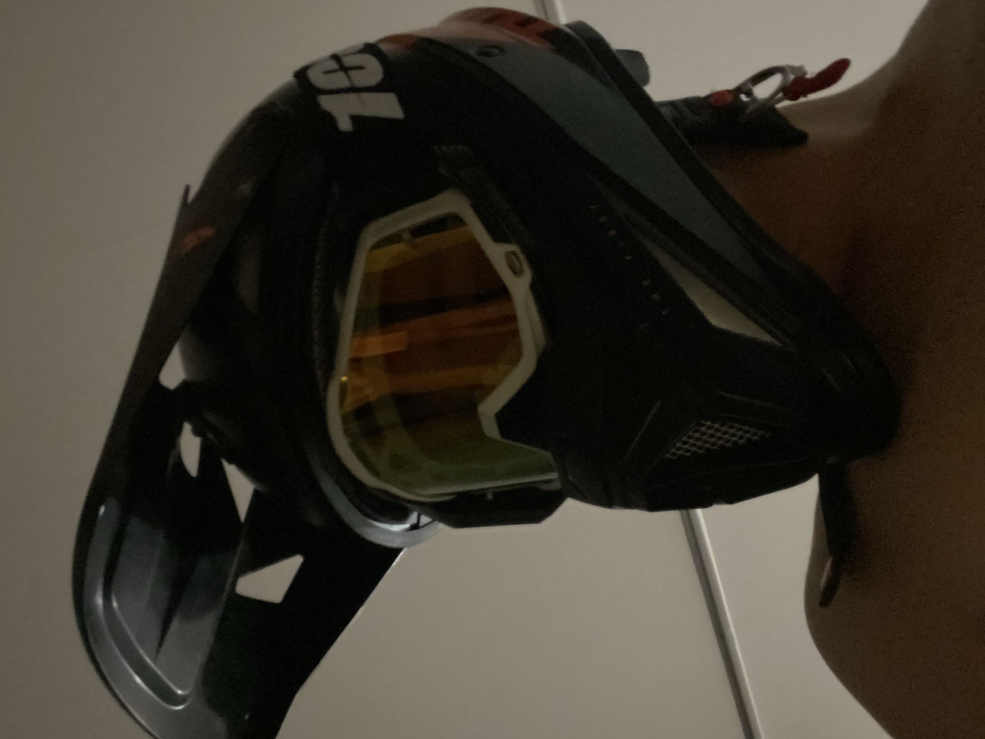 A close-up shot shows a black and orange full-face helmet with attached goggles, positioned near a person's shoulder. The helmet displays the number "100%", which is partially visible. The background features a plain wall.