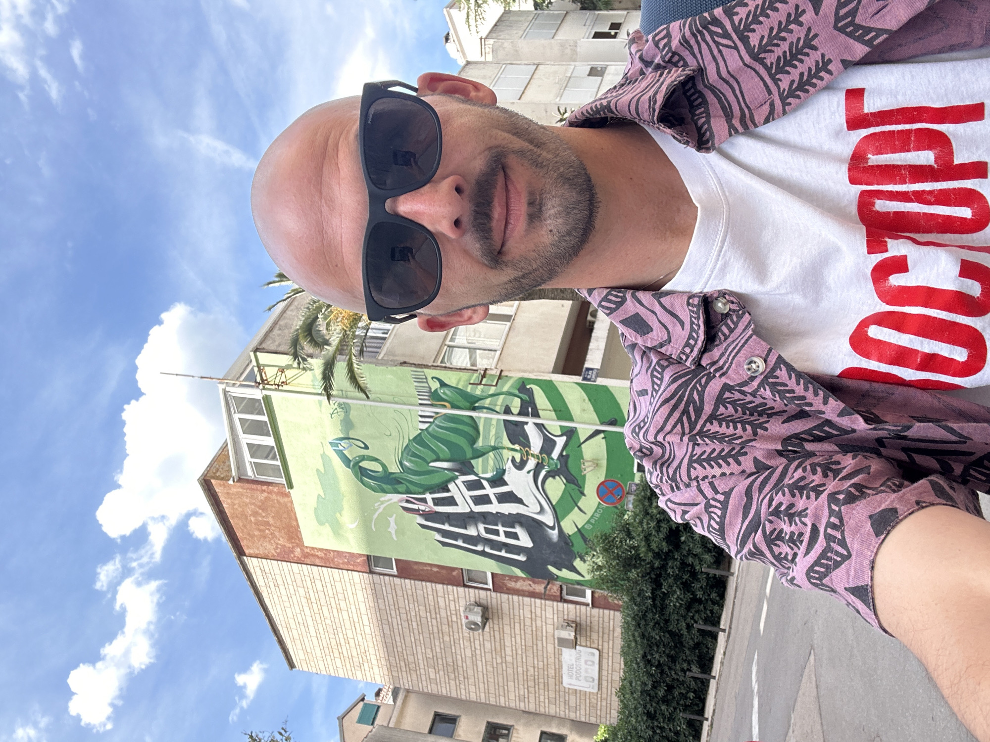 The image is a selfie of a bald man wearing sunglasses, a white t-shirt with a red inscription, and a pink patterned shirt. He is standing on a street next to a tall building. The building has a mural on its side depicting a green and black creature. The sky is blue with white clouds. The location is the city of Budva, Montenegro, judging by the text "Hotel Rodostomus" on the building.