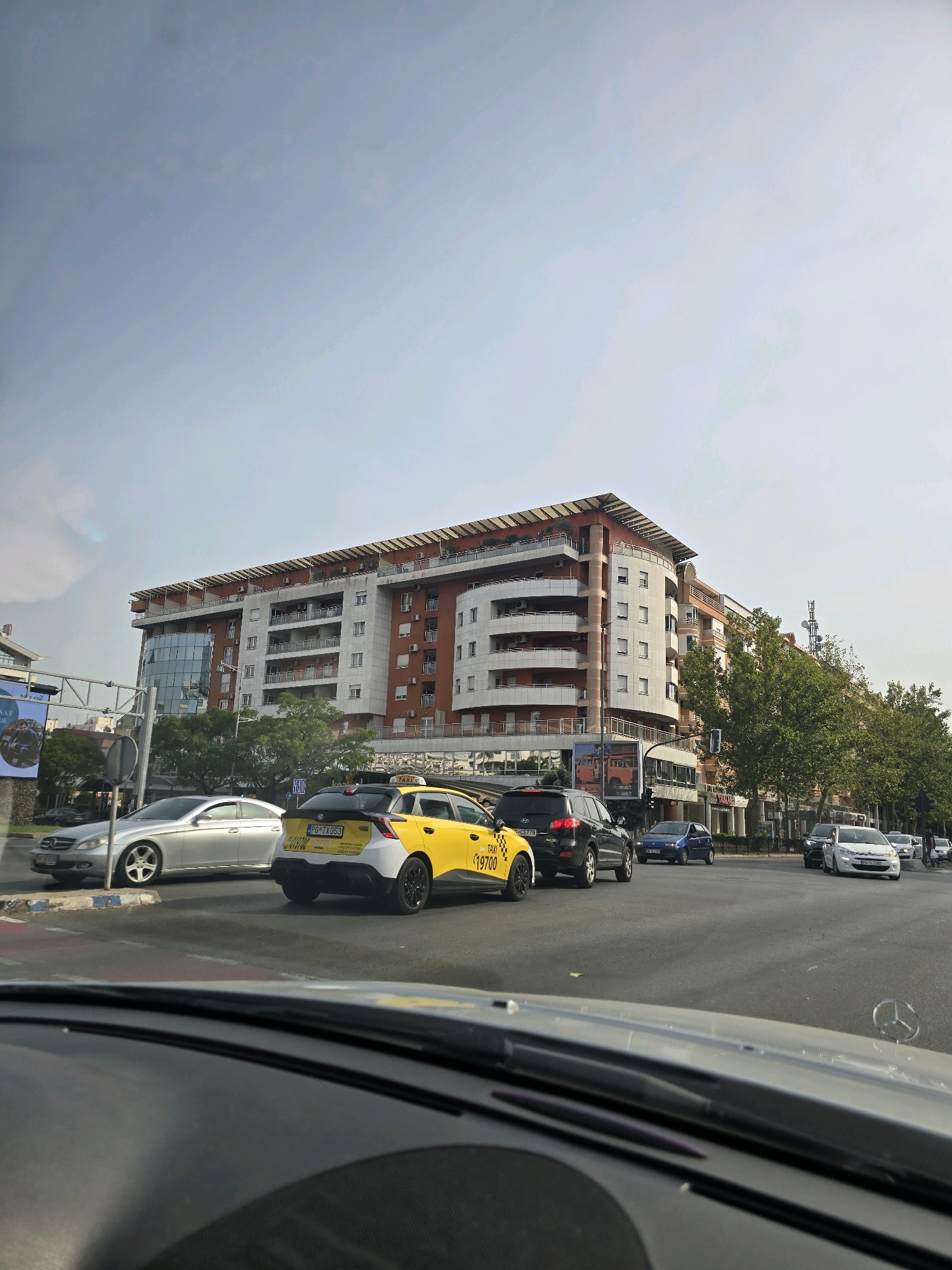 The image shows a view from inside a car, looking out at a city street in Podgorica, Montenegro. In the foreground, the dashboard of the car is visible, with a Mercedes-Benz logo. The street is busy with traffic. A yellow and white taxi is prominent in the center of the frame, with its license plate reading "PG TX G53". Other cars, including a silver Mercedes-Benz, a black SUV, and several white cars, are also on the road.
Behind the street, a large, modern building with a curved facade and balconies dominates the skyline. The building appears to be a residential or commercial complex, with several floors and a prominent red roof. Trees line the street in front of the building, and a billboard advertising something is partially visible on the left side of the image. The sky is overcast with a light blue hue. The scene appears to be captured during the daytime, likely late morning or early afternoon, based on the lighting conditions.