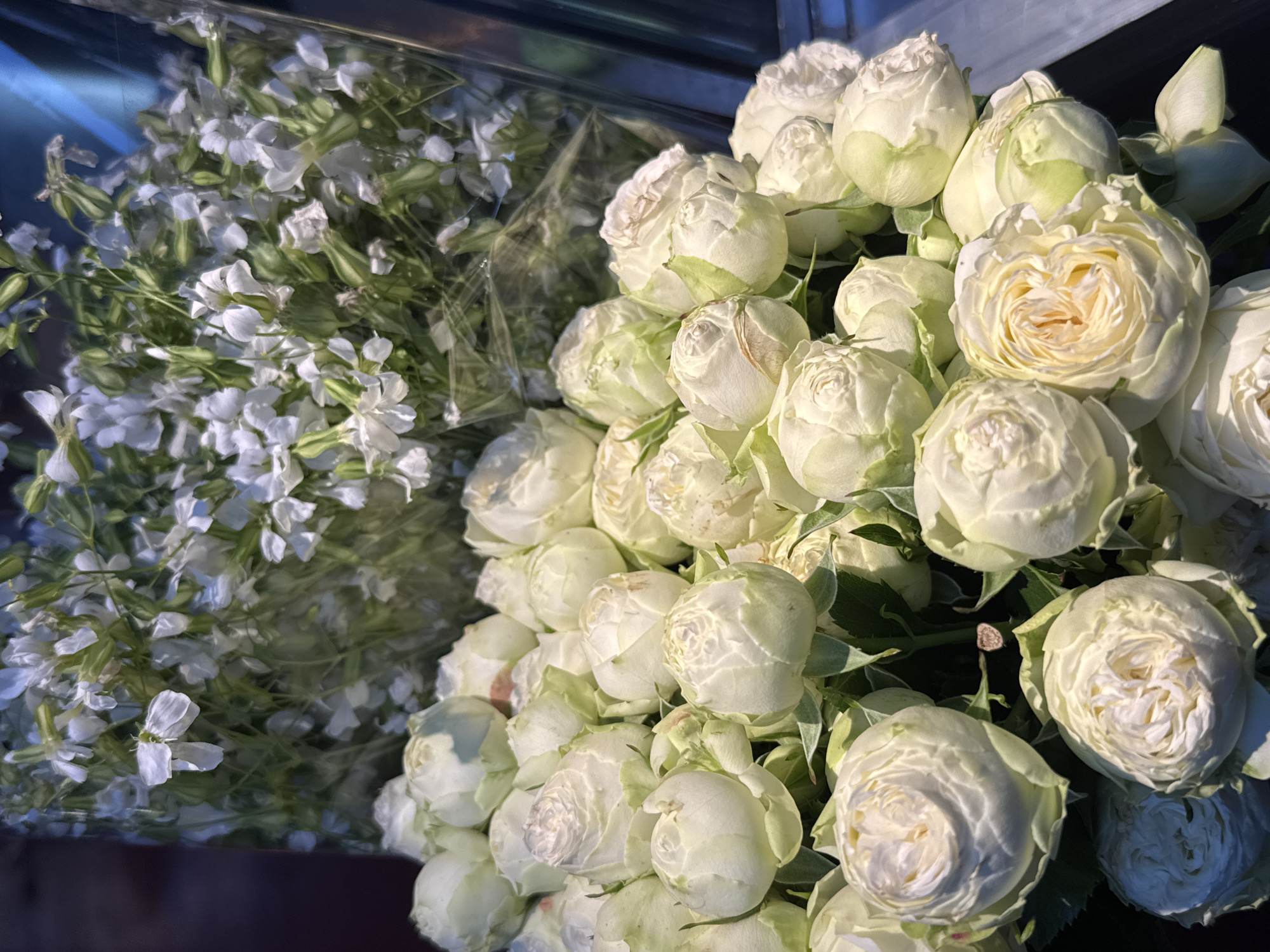 The image shows two bouquets of flowers. On the left, there is a bouquet of small, white, four-petaled flowers with visible green stems and foliage. The flowers appear delicate and densely packed. On the right, there is a cluster of white roses with a subtle hint of green on their outer petals. The roses are tightly budded, forming a round shape, and their petals are tightly packed. The lighting suggests an indoor environment, and it is unclear if there are any people present. The location of Yerevan, Armenia, cannot be determined from the image alone.