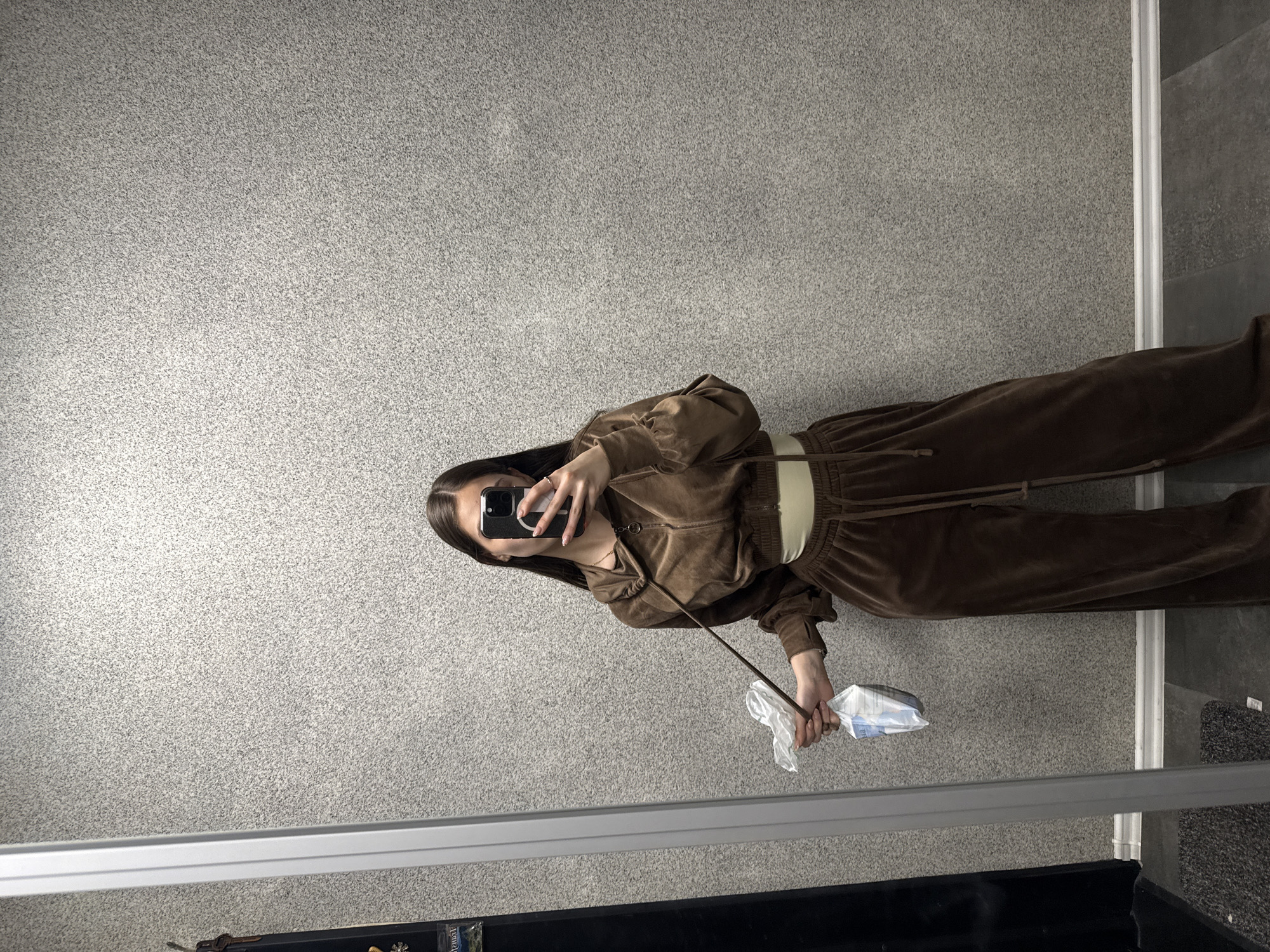 A young woman is standing indoors, likely in front of a mirror. She is dressed in a brown velour tracksuit and taking a selfie with her smartphone, which is obscuring her face. In her other hand, she holds a stick with a plastic-wrapped package attached. The background is a light gray carpet or wall. The lower portion of the mirror frame is visible, showing a black surface and a glimpse of a silver object. The image was likely taken in Sofia, Bulgaria, given the user’s location.