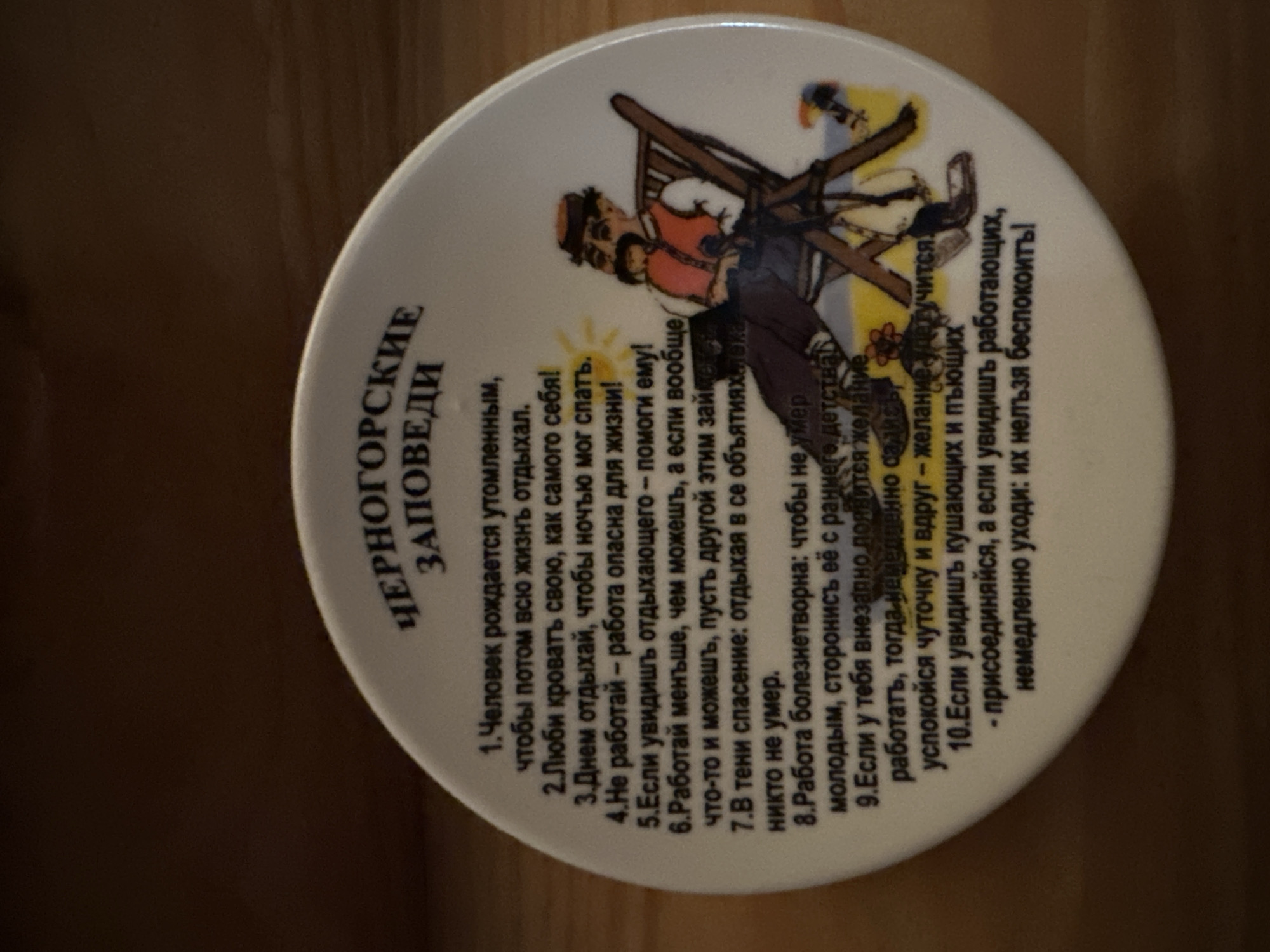 The image displays a white, round ceramic plate with text and an illustration. The text is in Russian and appears to be a list of humorous rules or advice. The title at the top reads "ЧЕРНОГОРСКИЕ ЗАПОВЕДИ" which translates to "MONTENEGRIN COMMANDMENTS". Below the title, there are numbered points, some of which are visible and partially legible, starting with "1. Человек рождается утомленным..." (1. A person is born tired...). An illustration features a cartoonish man with a mustache and hat, wearing a vest and sitting in a lounge chair on a yellow background, possibly representing a sunny day with a beach. The plate is angled and appears to be resting on a wooden surface. The lighting suggests indoor photography. There is no discernible connection to Sergeevka, Russia.