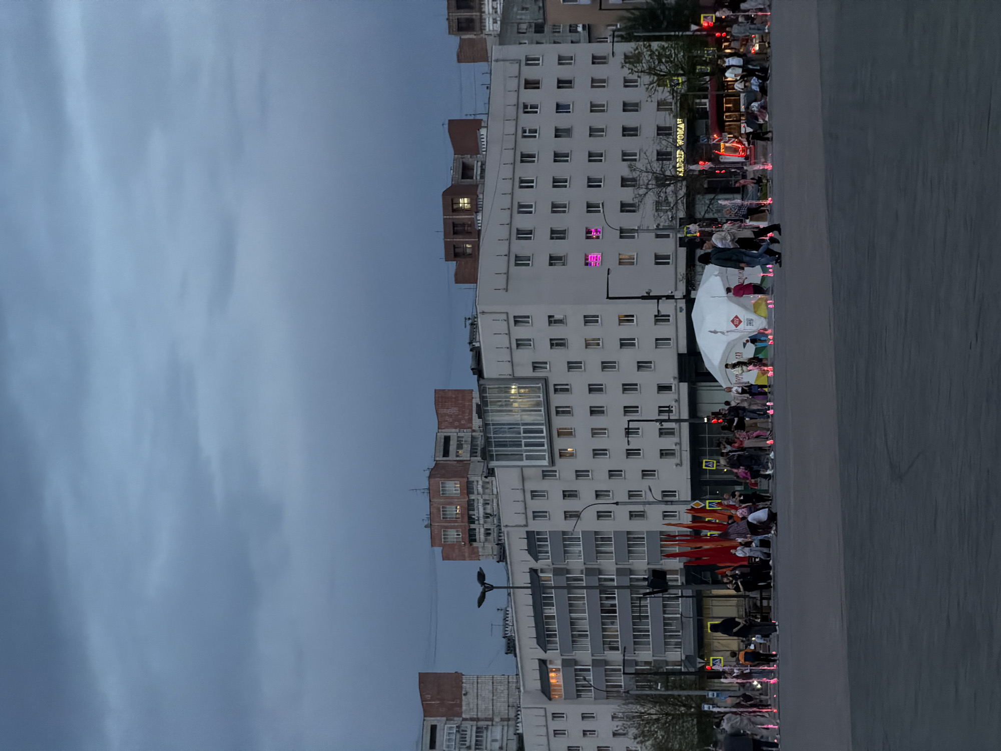 The image depicts a street scene in Perm, Russia, at dusk. Tall buildings line the street. A crowd of people is visible on the pavement, some are gathered near a white tent. Traffic lights are glowing red. A building has a sign in neon lights, and one of the windows is illuminated with pink light. The sky is cloudy.