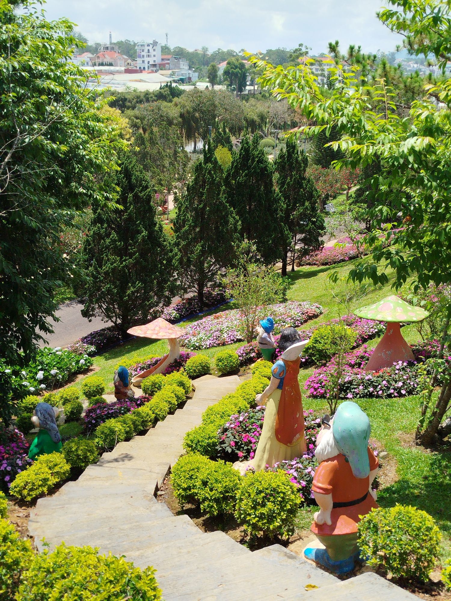 The image depicts a garden scene at the Da Lat Flower Gardens in Da Lat, Vietnam. A set of concrete steps descends into a garden area featuring statues of Snow White and the Seven Dwarfs. The garden contains various flowers and foliage, including rows of small, green shrubs and beds of pink and purple flowers. Concrete mushroom sculptures are also visible in the garden. In the background, a road is present, lined with more flowers and greenery. Beyond the garden, a cityscape with buildings and trees is visible under a cloudy sky. The lighting suggests a bright, sunny day.