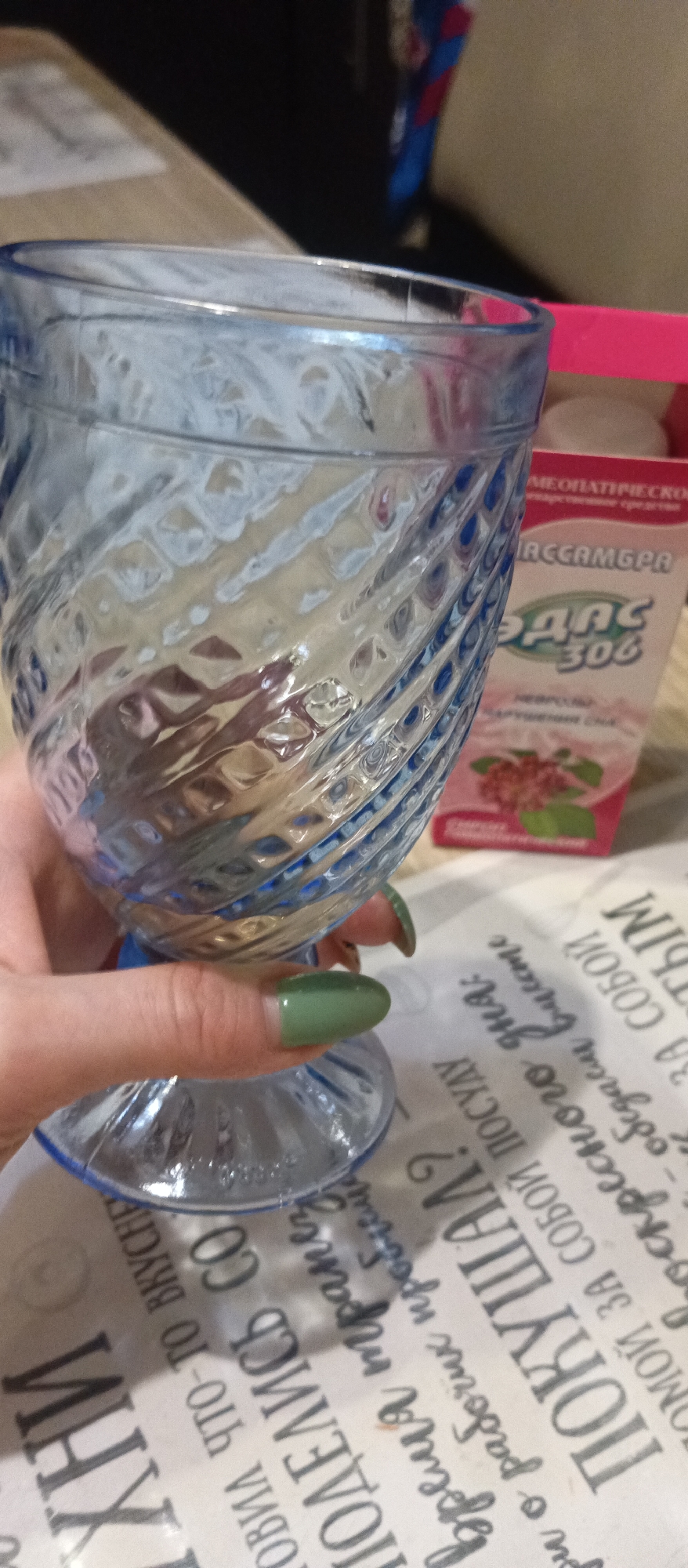 A person with green nail polish is holding a blue glass with a textured swirl pattern. Next to the glass is a pink and white box labeled "ЭДАС 306", which appears to be a homeopathic medicine. The glass rests on a white surface with black text in Russian. The words "ПОКУШАЛ?" are prominently visible on the surface. A blurred box sits in the background. The location is presumably indoors.