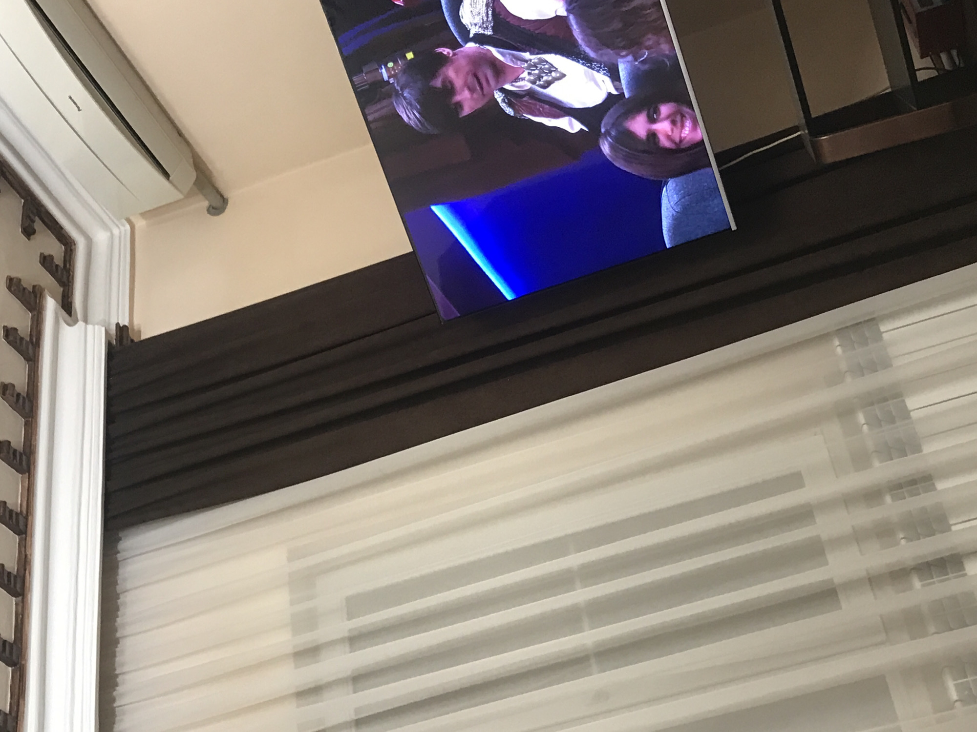 A tilted indoor shot captures a television screen displaying two people, one with short brown hair and the other with dark hair, set against a blue-lit background. The television is positioned above brown drapes and a window with white blinds. A white air conditioning unit is mounted on the wall to the left, adjacent to a decorative molding. The location is suggested to be Yerevan, Armenia.