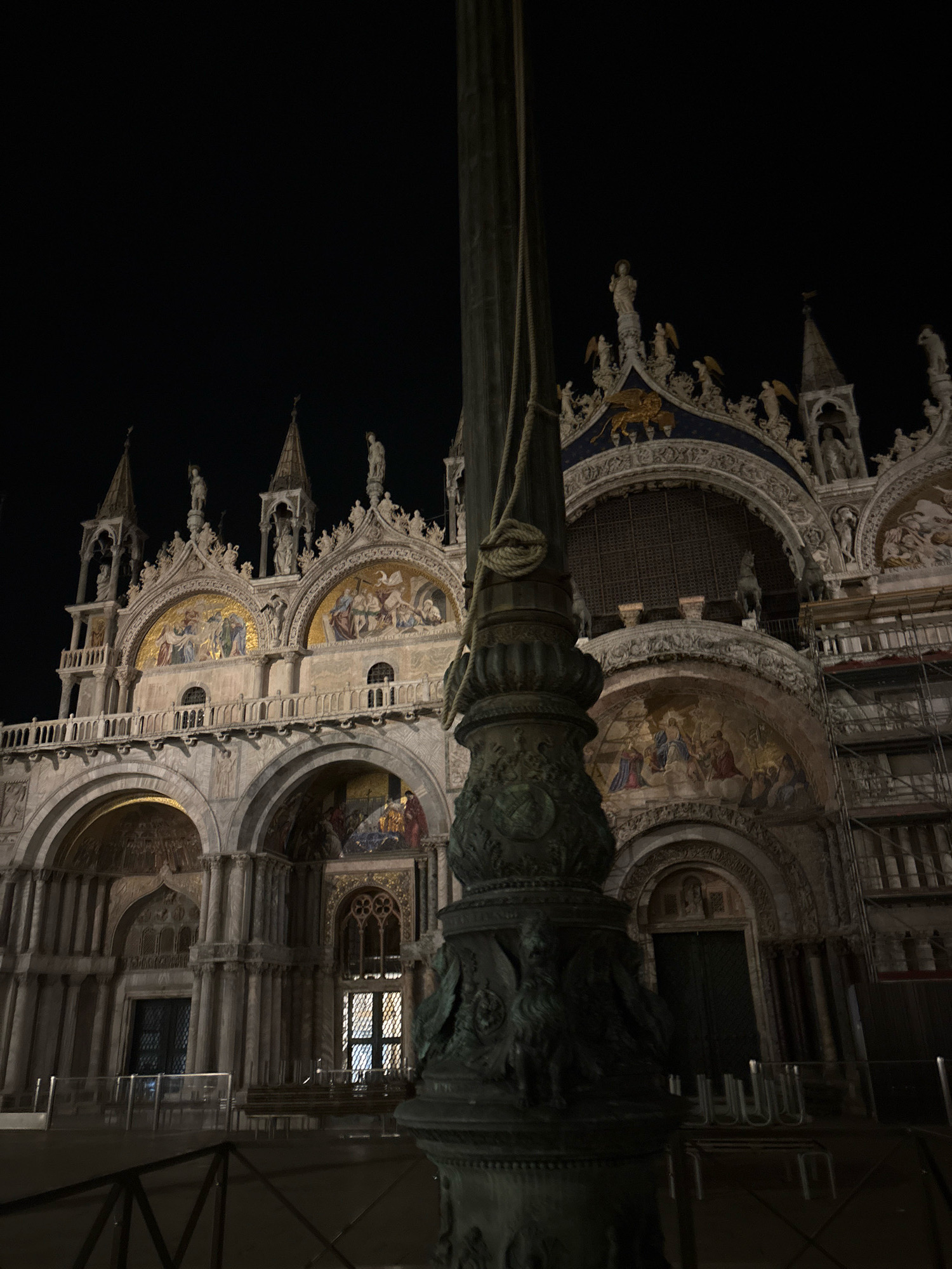 The image shows a night-time view of St. Mark's Basilica in Venice, Italy, with a large lamppost in the foreground. The basilica, a Roman Catholic cathedral, is illuminated, highlighting its intricate facade with arches, mosaics, and sculptures. The sky above is dark, indicating it is nighttime. The lamppost is dark and ornate, with a rope tied around its middle. In front of the basilica, there are railings and benches, suggesting it is a public space. The overall scene is grand and historical, typical of Venetian architecture.
