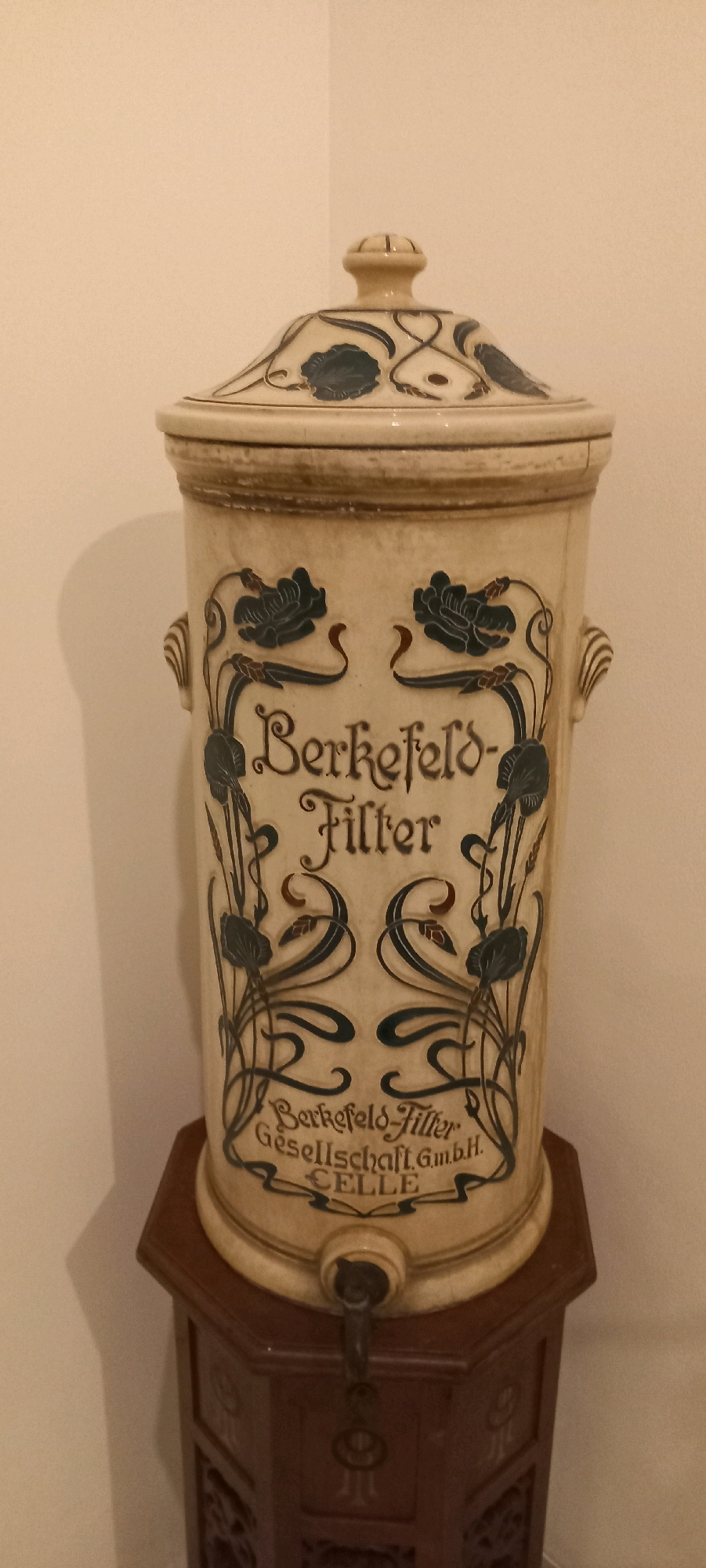 The image shows a cream-colored ceramic water filter with dark blue floral decorations and text. The text on the filter reads "Berkefeld-Filter Gesellschaft G.m.b.H. CELLE". The filter is cylindrical with handles on the sides and a domed lid. A tap is visible at the bottom of the filter. The filter is placed on a dark brown, octagonal wooden stand with intricate carvings. The background is a plain, light-colored wall, with a shadow cast on it from the filter. The scene appears to be an indoor setting. The provided information about the location in Central Jakarta, Indonesia is not supported by any visual cues in the image.