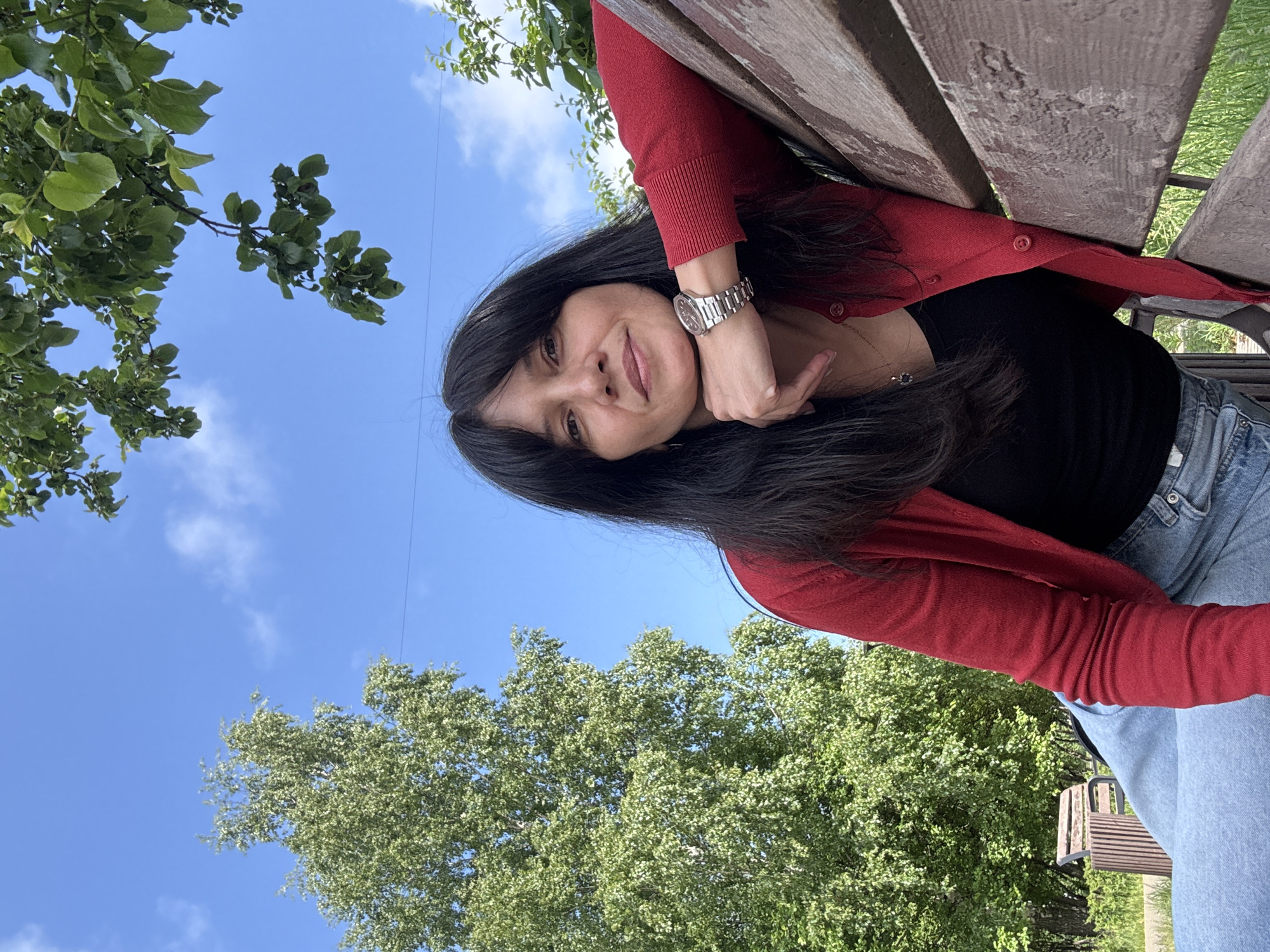 The image captures a young woman outdoors in what appears to be a park or green area in Chaikovsky, Russia. She is positioned on her side, leaning against a wooden bench, with her head resting on her hand. She has long, dark hair and is wearing a red cardigan over a black top and light blue jeans. A silver-toned watch is visible on her left wrist, and she wears a delicate necklace. She looks directly at the camera with a calm, pleasant expression and a slight smile.
The scene is set under a bright blue sky with scattered white clouds. Lush green trees with full foliage frame the upper and lower portions of the image, suggesting a spring or summer day. The photograph is taken from a low angle, looking upwards, and is rotated 90 degrees clockwise, giving a unique perspective of the woman against the sky and trees. No visible text is present in the image.