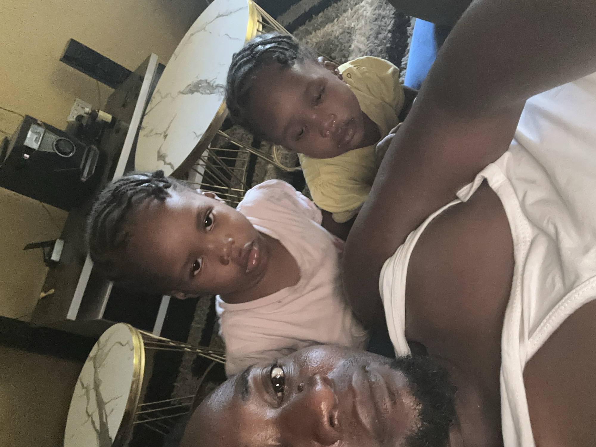 The image shows a man and two young children, possibly in a home setting. The man, who appears to be taking a selfie, is positioned at the bottom of the frame. One child is looking towards the camera wearing a light pink shirt, while the other child is wearing a yellow shirt and appears to be sleeping or resting. A table with a marble-like surface and gold trim is partially visible in the background. There is also a stereo visible, with a rug on the floor. The city and location are unknown.