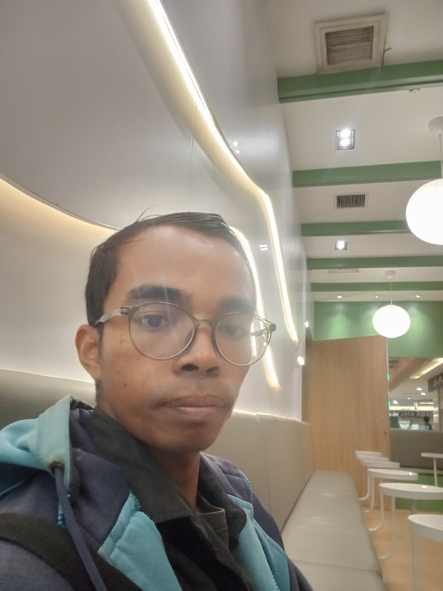 A man with glasses is taking a selfie indoors, possibly in a cafe or waiting area. He is wearing a dark blue and light blue hooded jacket. The background shows a modern interior with white curved walls, soft lighting, and green accents. There are rows of white stools and tables in the distance, and a wooden panel wall. A sign with Indonesian text, possibly "JAYA ABADI," is visible in the background. The lighting suggests it could be a shopping mall or a public space in Surabaya, Indonesia, given the signage.