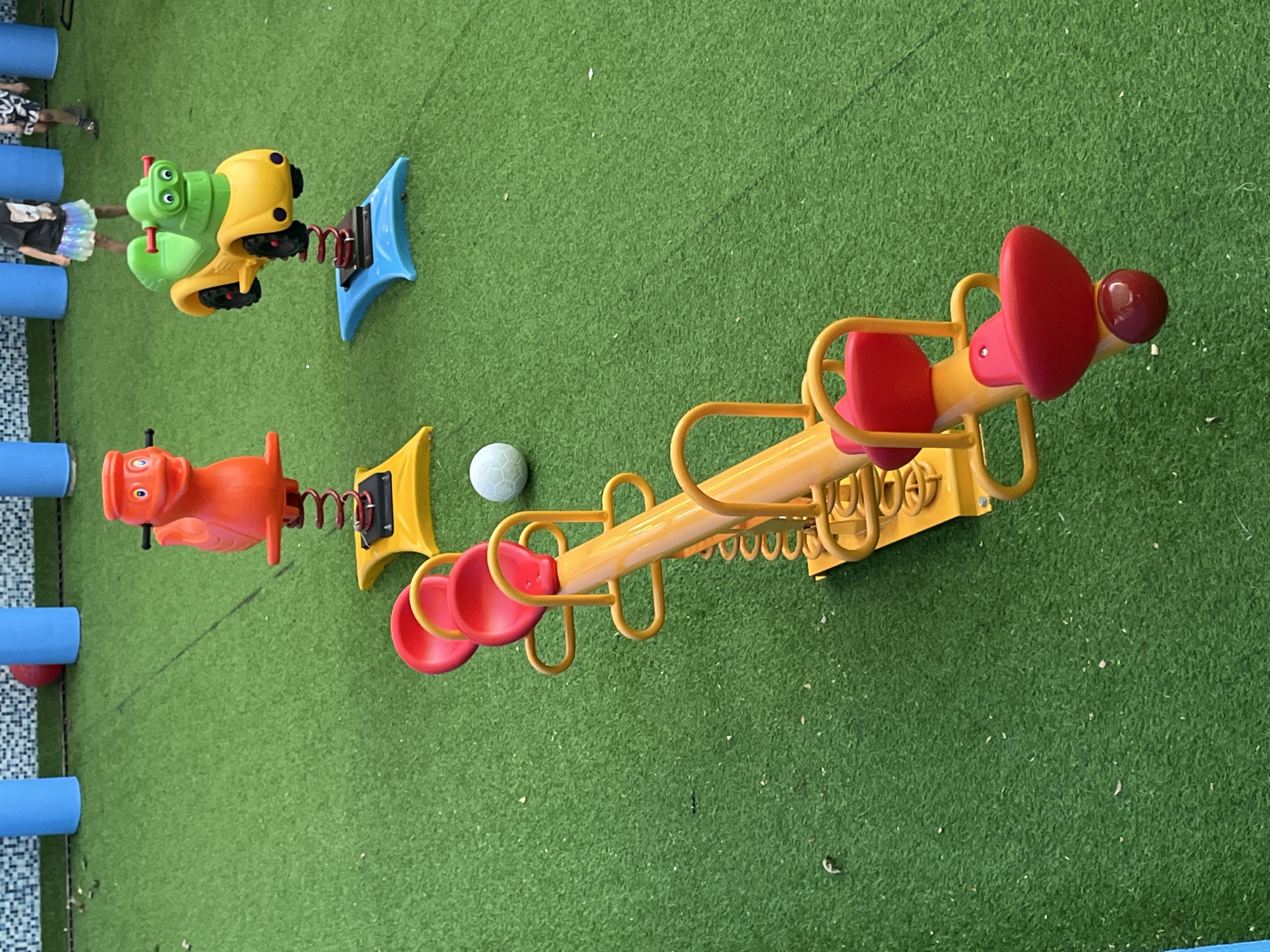 The image shows a playground with artificial turf in Nha Trang Ward, Vietnam. In the foreground, a yellow seesaw with red seats is positioned diagonally across the frame. Further back, there are two spring-rider toys: one is an orange dinosaur and the other is a yellow and green vehicle resembling a car or motorcycle. In the background, out-of-focus, are blue cylindrical structures and two children. One child is wearing a black t-shirt and a colorful skirt, and the other is wearing shorts. A red ball is partially visible behind the blue structures.