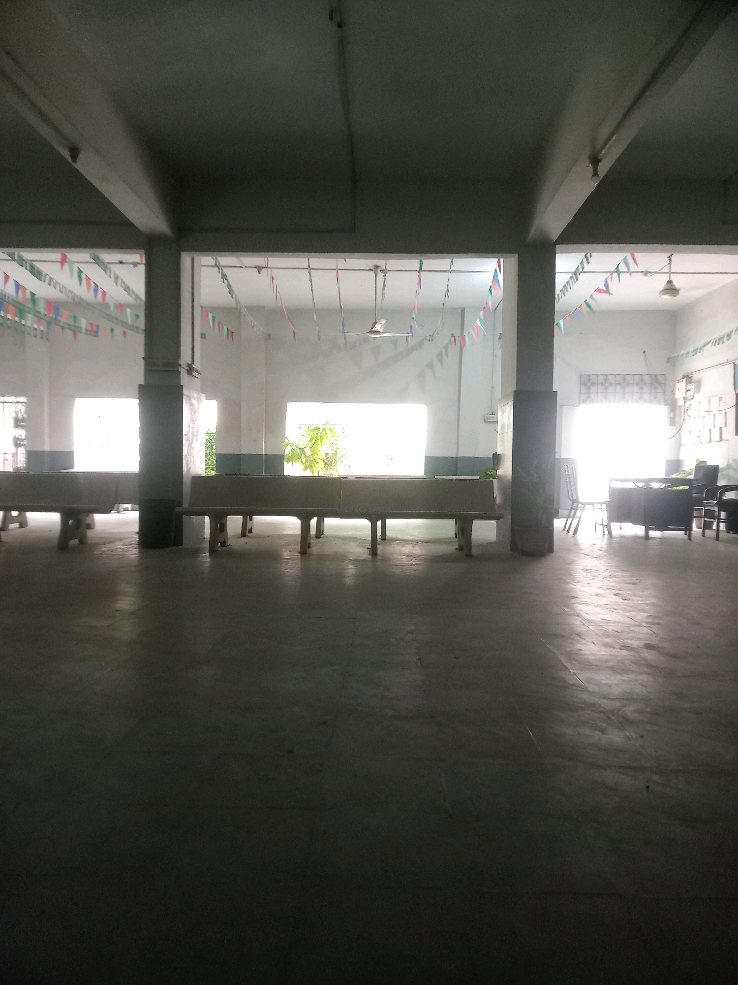 The image depicts a brightly lit, covered outdoor area or hallway, likely within an institution like a school or government building, given the presence of flags and what appears to be a reception desk. The space is characterized by concrete floors and pillars supporting a ceiling. Along the walls, large windows or open archways reveal more of the interior or possibly another section of the building. Strings of triangular flags in various colors are strung across the upper portion of the space. In the foreground, there are long, concrete benches on either side, suggesting a waiting or seating area. Towards the back of the image, a reception desk with chairs and office equipment is visible. The overall impression is one of a public or communal space, possibly in a school setting. Without specific identifying markers, pinpointing the exact location in Karachi Division is not possible.