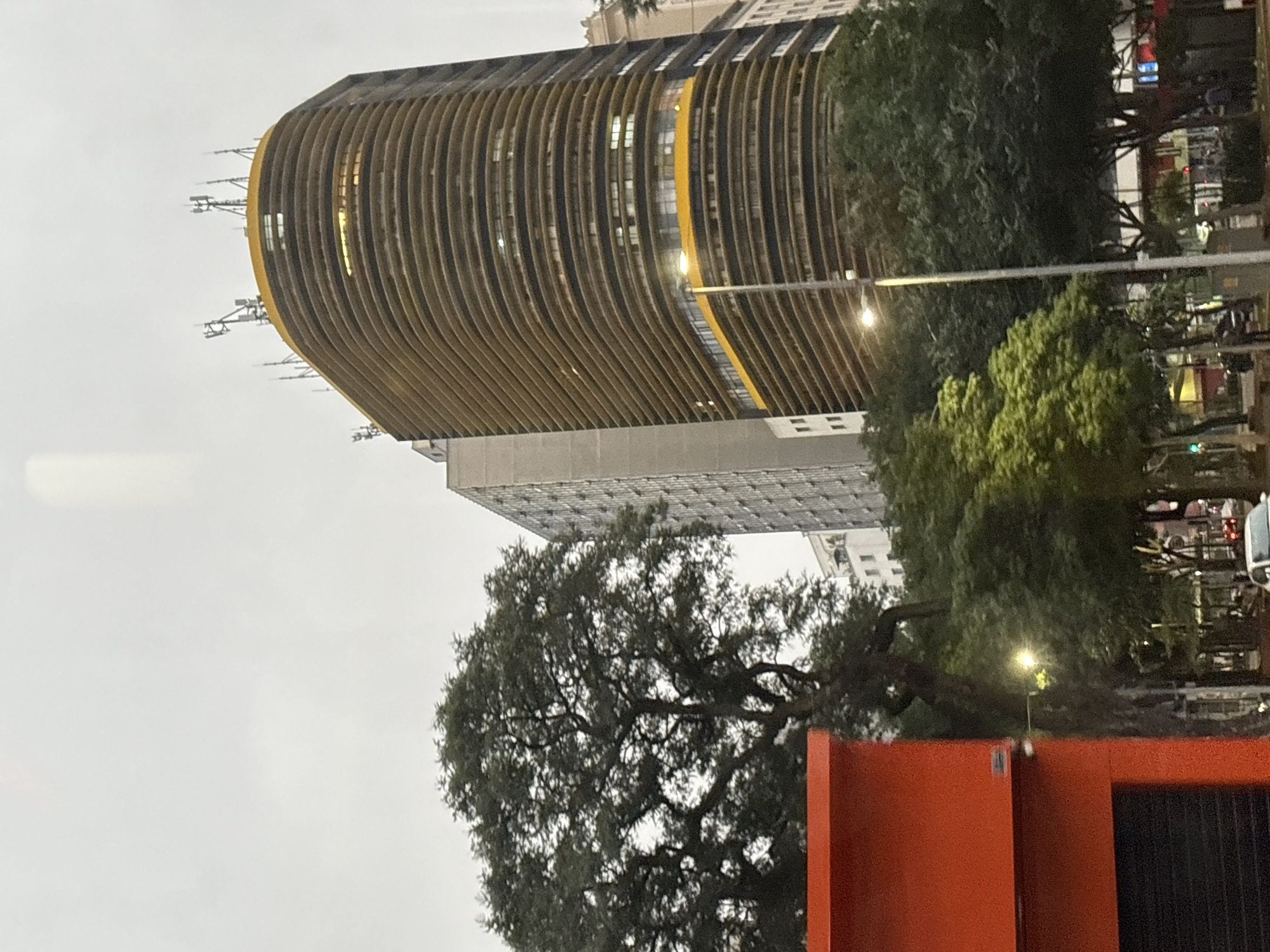 This image captures a street view in São Paulo, Brazil, featuring the iconic Copan Building and neighboring structures under an overcast sky. In the foreground, a vibrant orange structure with a black roller-shutter door occupies the left side. Dominating the mid-ground is the distinctive curved facade of the Copan Building, characterized by its horizontal banding and a bright yellow accent at the top and middle sections. Numerous antennae are visible on the building's roof. To the left of the Copan Building, a tall, rectangular skyscraper with a grid-like pattern of windows stands. Several large trees with dense foliage are interspersed throughout the scene, with their branches reaching towards the sky. Streetlights and traffic signals are also present, hinting at an urban environment. In the distance, other buildings and vehicles are partially visible, suggesting a busy city street. The overall impression is one of a densely built urban landscape on a cloudy day.