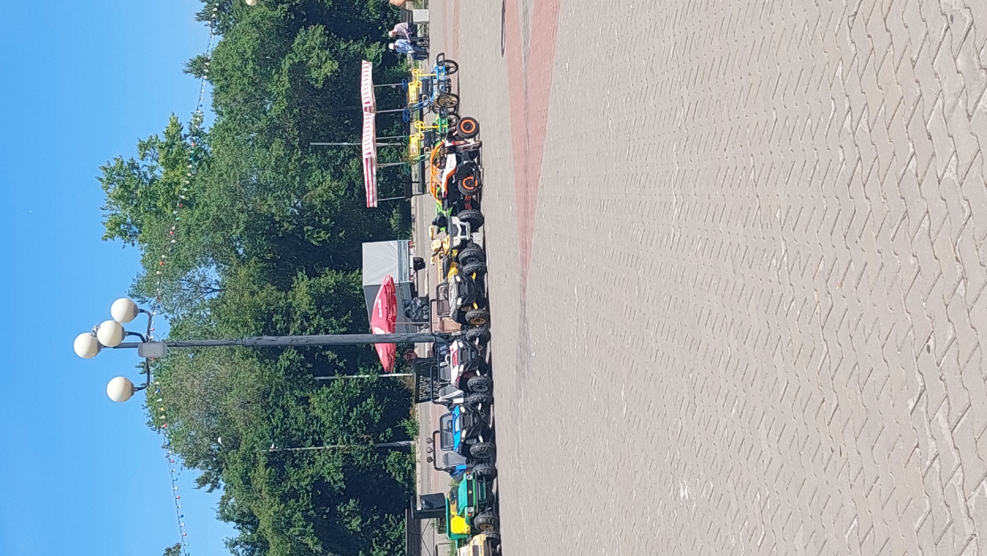 The image shows a sunny day in a city park in Komsomolsk-on-Amur, Russia. A paved area, likely a promenade or square, is in the foreground, with patterned grey brickwork. In the middle ground, a row of child-sized electric vehicles, resembling small ATVs and buggies, are parked. They are various colors, including green, blue, white, black, and orange. Behind the vehicles, a red umbrella shades a table and chairs. Further back, a white trailer is parked, and behind that, a red and white striped canopy covers seating areas. In the distance, a line of lush green trees forms the edge of the park. A tall lamppost with multiple light fixtures stands prominently on the left side of the image. Several people are visible: two individuals are seated on a bench in the background, and another person appears to be standing near the red umbrella. The sky is clear and blue, suggesting good weather.
