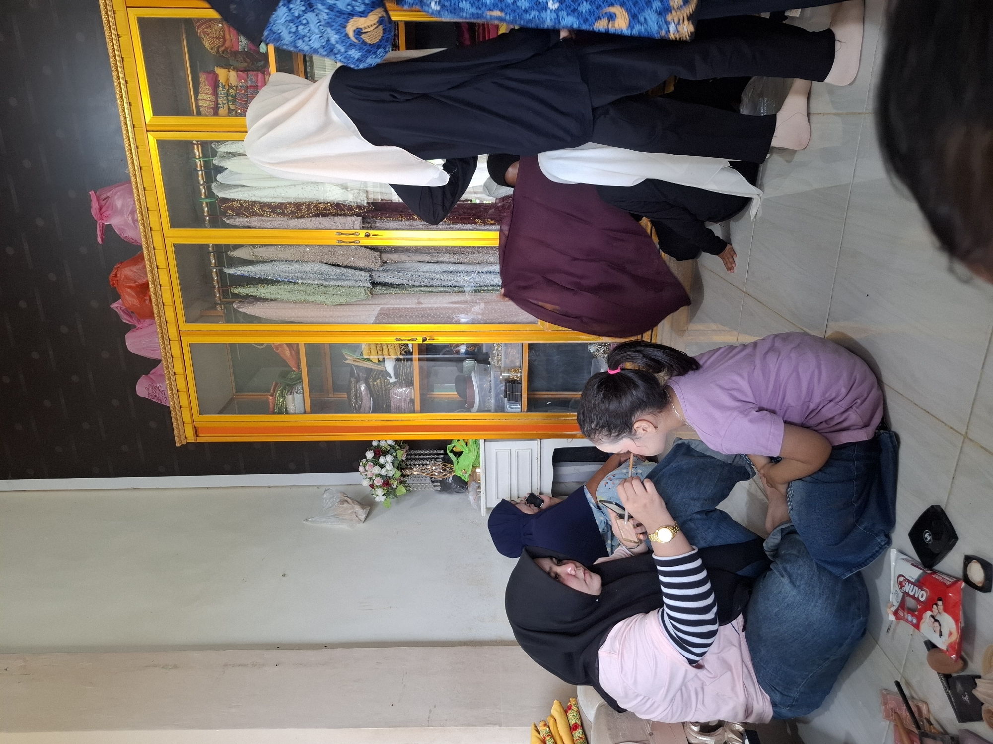 In Beurawang, Indonesia, a scene unfolds indoors with a focus on personal grooming. A woman, wearing a black hijab and a pink striped shirt, is applying makeup to a young girl who is wearing a purple shirt. The girl is sitting cross-legged, and the woman is gently touching her face with a makeup tool.

To the left, a small beige sofa is visible, with yellow fabric stacked on it. A white cabinet with drawers stands behind the woman and girl, adorned with a floral arrangement in a decorative vase.

In the background, a prominent tall yellow cabinet with glass doors displays an assortment of colorful garments on hangers and neatly folded fabrics. Pink plastic bags are perched on top of this cabinet.

Several other individuals are present in the scene. A person in a blue hijab is seated behind the makeup artist, appearing to be on a phone call. Another person in a white hijab and black clothing is seen partially obscured, kneeling. Further to the right, a person wearing a blue and gold patterned garment is visible, as is another person in black attire. The floor is tiled with large square tiles. Various makeup items, including compacts and brushes, are scattered on the floor in the foreground. The lighting suggests an indoor environment, likely a home or a small shop.