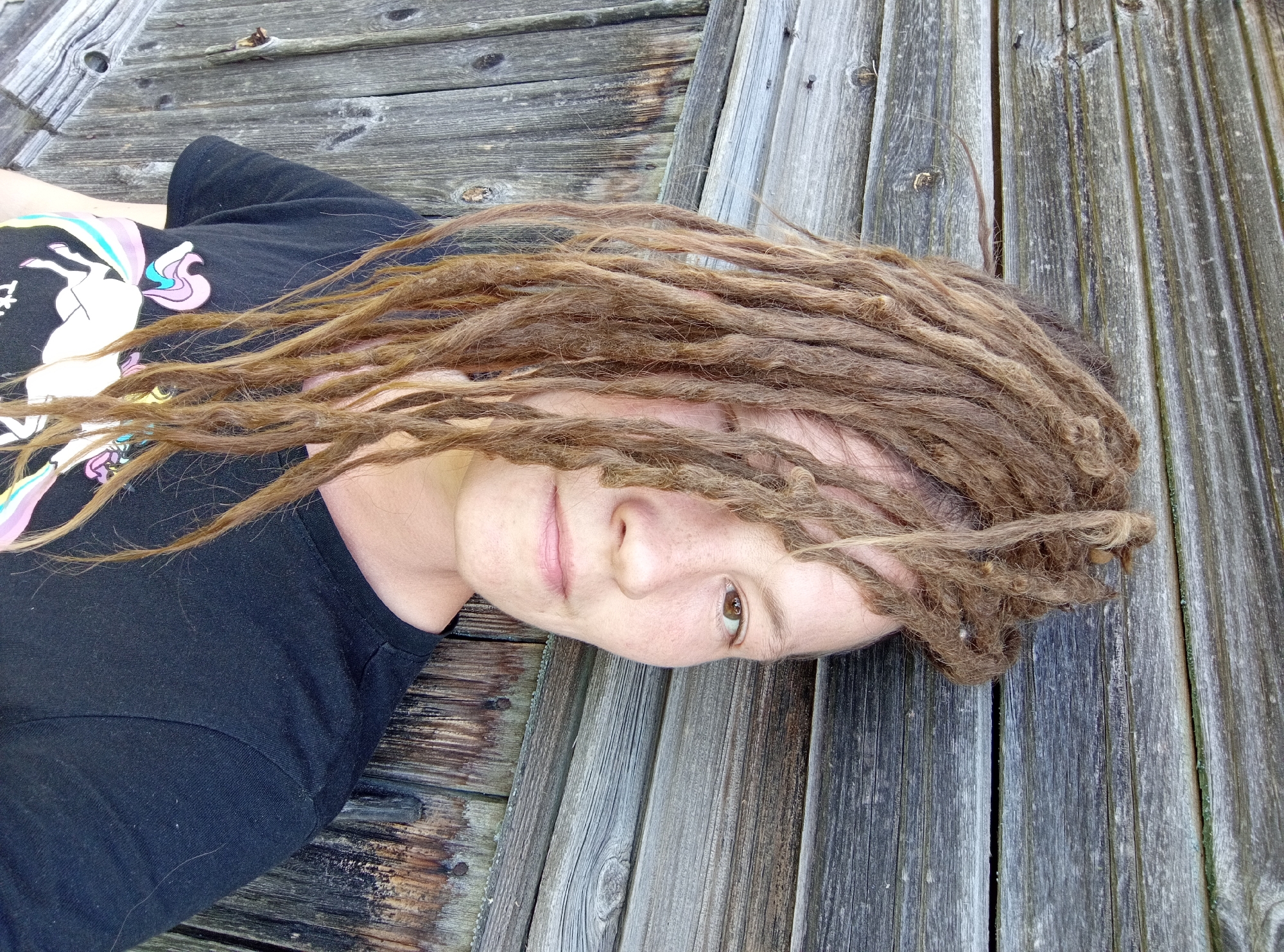 A person with light brown dreadlocks is captured in a close-up, angled shot, lying outdoors on a weathered wooden surface. The individual is wearing a black t-shirt adorned with a colorful unicorn graphic. Their dreadlocks are spread around their head, partially covering the left side of their face, but their right eye, nose, and mouth are clearly visible. They display a relaxed expression, with a subtle smile and their right eye looking directly at the camera.
The setting appears to be a wooden deck or similar structure, characterized by aged, greyish planks with prominent grain, knots, and visible gaps. The natural lighting suggests it is daytime. The scene evokes a calm and informal atmosphere. The location context for this image is Ostrovtsy, Russia. No other discernible text is visible within the image beyond the unicorn design on the t-shirt.