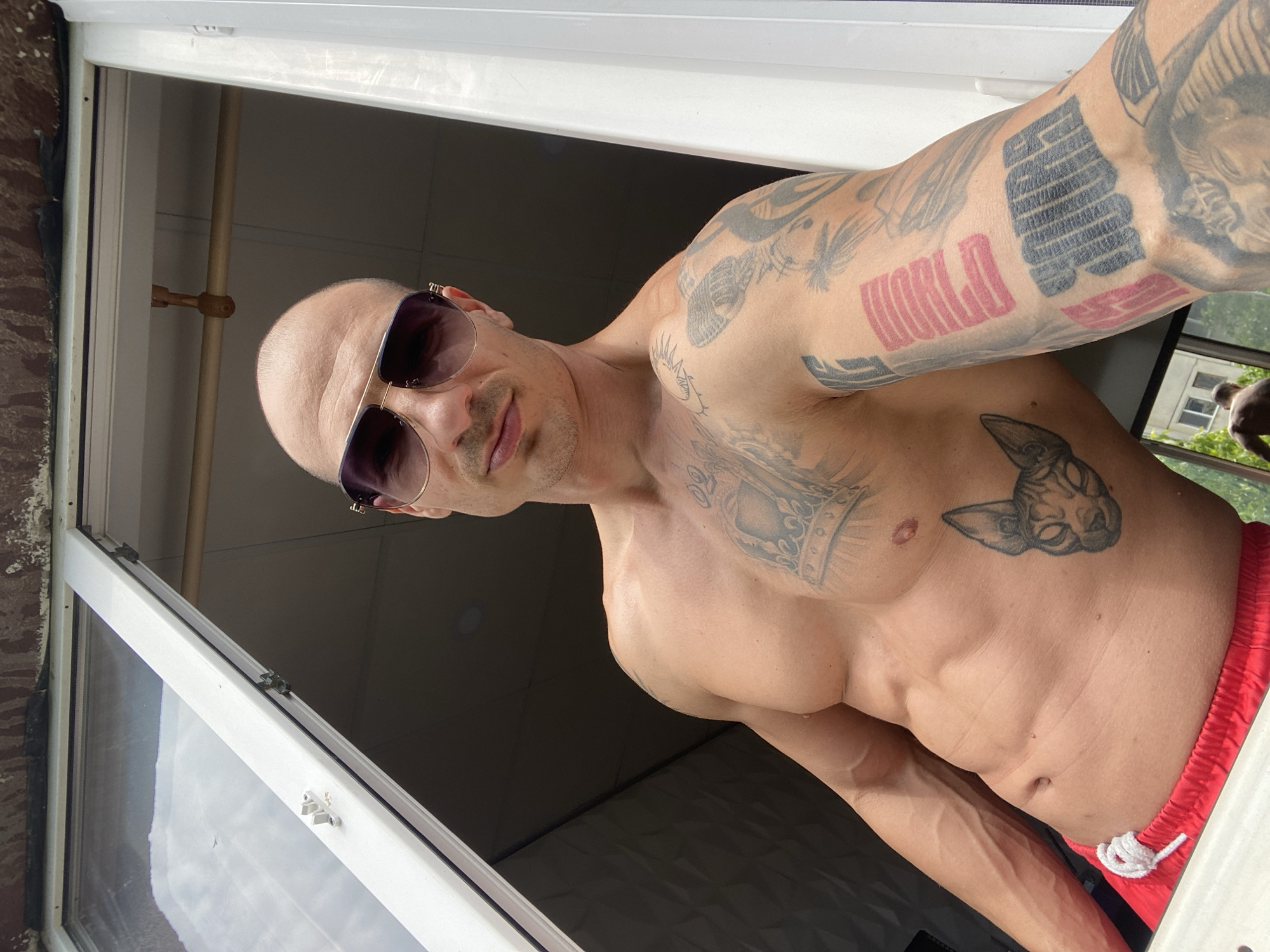 A bald man wearing sunglasses and red shorts is posing in a room with an open window. The man is shirtless and has tattoos on his torso and arms. He is positioned at an angle, looking towards the camera. The location of the image is unknown, and there is no way to prove this image was taken in Pleven, Bulgaria.