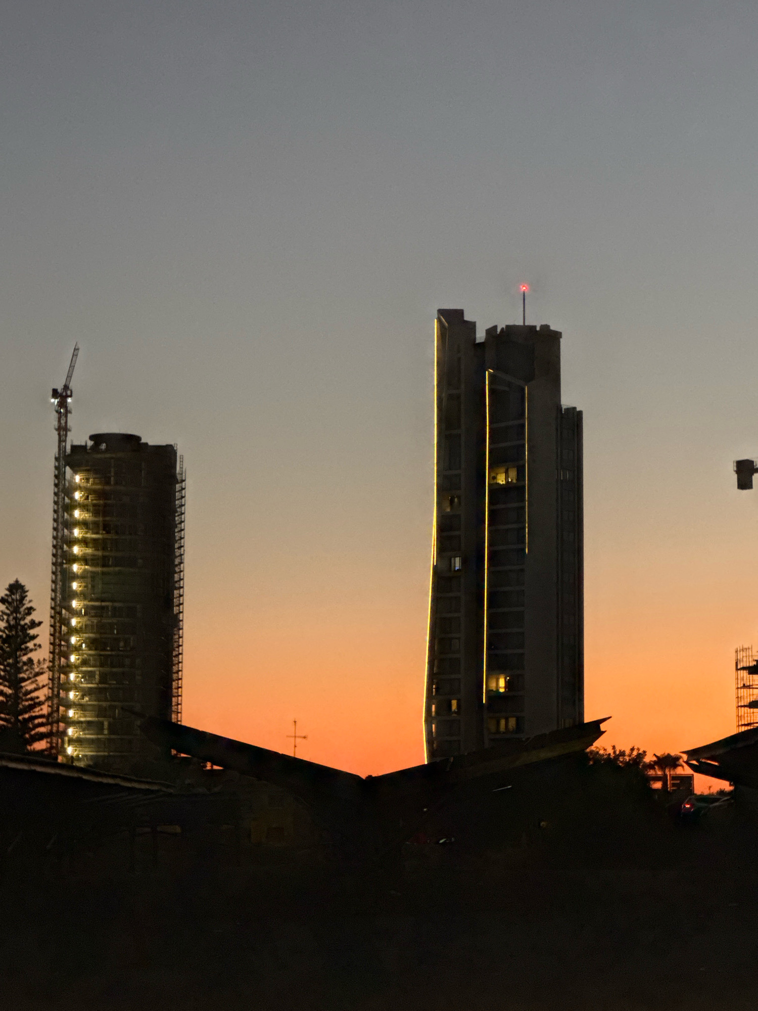 An outdoor, low-angle, medium shot captures a twilight cityscape scene in Mouttagiaka Community, Cyprus. In the foreground, the dark silhouettes of rooftops and what appear to be construction materials are visible against an orange-grey gradient sky. Two prominent high-rise buildings dominate the mid-ground. The building on the left is a cylindrical skyscraper under construction, wrapped in scaffolding and illuminated by rows of yellow lights. A construction crane is attached to its upper section. The building on the right is a taller, more angular skyscraper with vertical yellow lighting accentuating its facade. This building has a small red light at its peak. In the far right, a dark, angular structure, possibly another building under construction or equipment, is partially visible. The overall atmosphere is one of urban development during a transitional time of day.