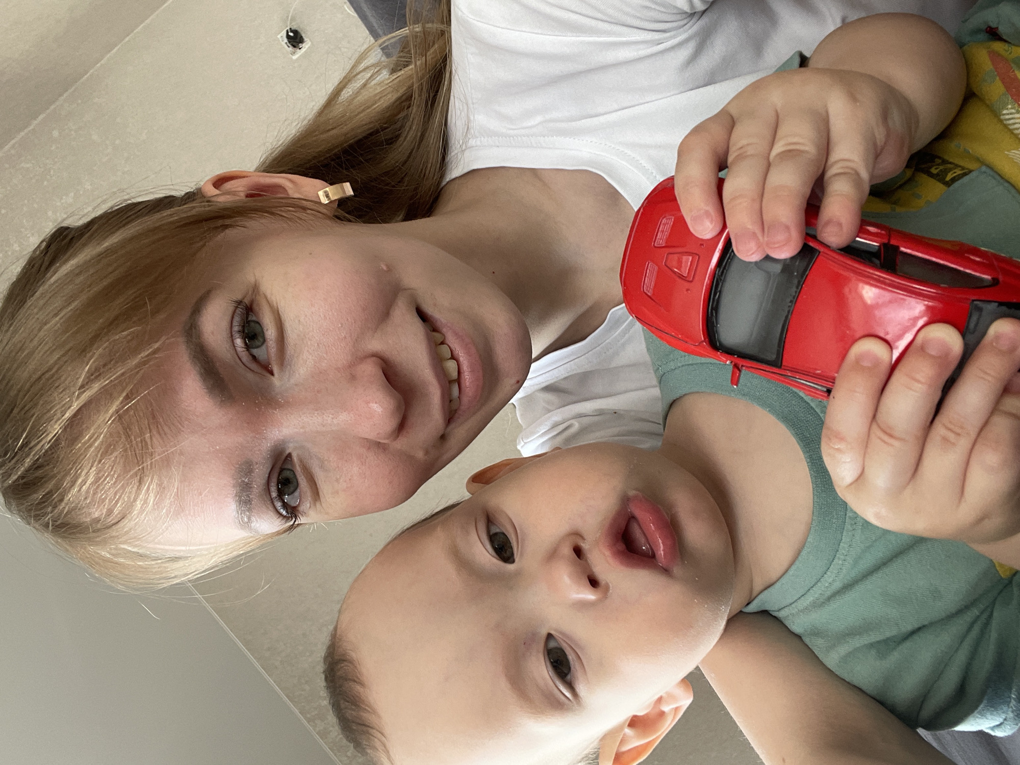A Caucasian woman and a young child are depicted in a selfie. The woman is fair-skinned with blonde hair, and her ear is visible, adorned with a rectangular earring. She is wearing a white t-shirt and smiling. The child, whose face is angled slightly downward, has fair skin and is wearing a green shirt. The child's mouth is slightly open. They both hold a red toy car. The background is a light, smooth surface, possibly a wall or floor. The location is noted to be Perm, Russia, but there are no visual indicators in the image to confirm this.
