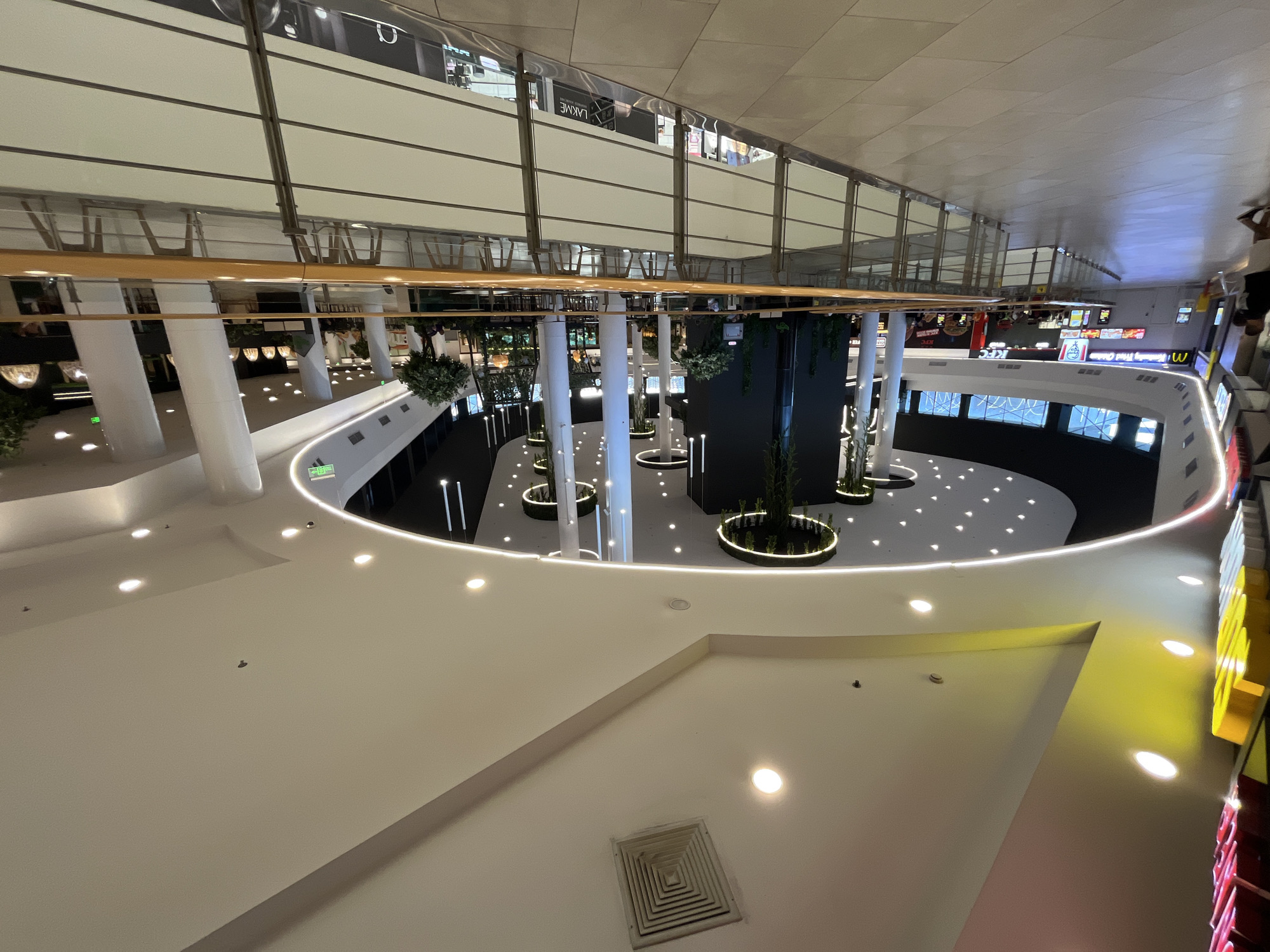 The image depicts the interior of a modern shopping mall in Stara Zagora, Bulgaria. The perspective is from an elevated walkway, looking towards a food court area. The mall features a multi-level design with a central atrium.
Dominant architectural elements include a curved ceiling with recessed lighting, circular decorative light fixtures suspended from the ceiling, and large white cylindrical pillars supporting the upper levels. A polished tiled floor is visible in the foreground and on the walkway. A metal railing with a wooden top separates the walkway from the lower level.
In the food court area, there are tables and chairs arranged for dining. Various fast-food establishments are visible, including McDonald's and KFC, indicated by their signage. Artificial trees and greenery are used as decorative elements throughout the mall.
The lighting is a mix of ambient overhead lighting and accent lighting, creating a bright and airy atmosphere. The overall impression is that of a contemporary and spacious shopping environment.