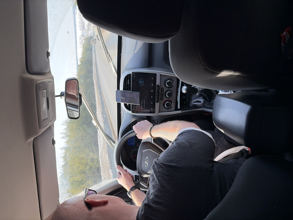The scene is captured from the interior of a vehicle, looking forward from a rear passenger seat on a sunny day in Zabegalovo, Russia. A bald, male driver, wearing sunglasses and a dark short-sleeved shirt with white trim, operates the car, his hands positioned on the steering wheel. A black digital watch is on his left wrist and a dark beaded bracelet on his right. The car's dashboard features a central infotainment screen, climate controls, and a smartphone mounted vertically above the central vents, displaying a navigation application. The instrument cluster shows the vehicle speed at approximately 60-70 units (likely km/h). A manual transmission gear stick is visible in the center console. The windshield is noticeably dirty with streaks, and the wipers are at rest at the bottom. Through the windshield, a two-lane paved road is visible, curving to the right. The road shoulders are partially covered in snow. A dense evergreen forest lines the left side of the road. In the distance on the right, a snowy, open landscape with scattered buildings or a settlement is discernible under a clear sky. A reflection, likely of the driver's head, is visible in the rearview mirror. A beige seatbelt buckle is seen unbuckled between the driver's seat and the center console armrest.