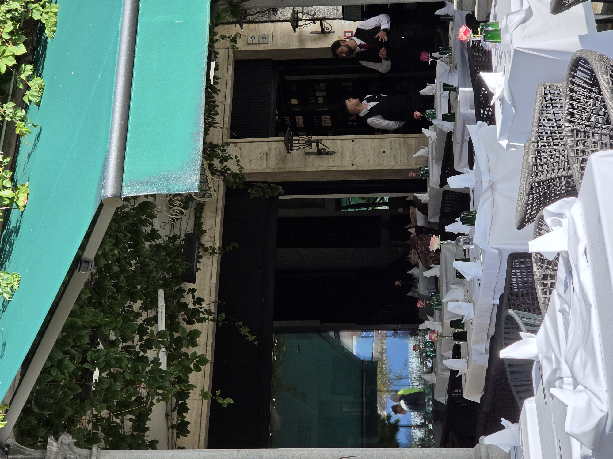 This image captures an outdoor dining area of a restaurant in San Giovanni, Italy, under a bright green awning. The tables are meticulously set with white tablecloths, folded white napkins, and small pink roses in green vases, suggesting an elegant and well-maintained establishment. Two male servers, dressed in black vests over white shirts, are visible near the entrance of the restaurant, engaged in conversation. One server is holding a tablet or menu. In the background, through a large glass window, a reflection shows people and trees, hinting at the surrounding environment. The architecture features stone pillars and a building with an ornate facade. The overall scene conveys a sense of sophistication and pleasant dining ambiance.
