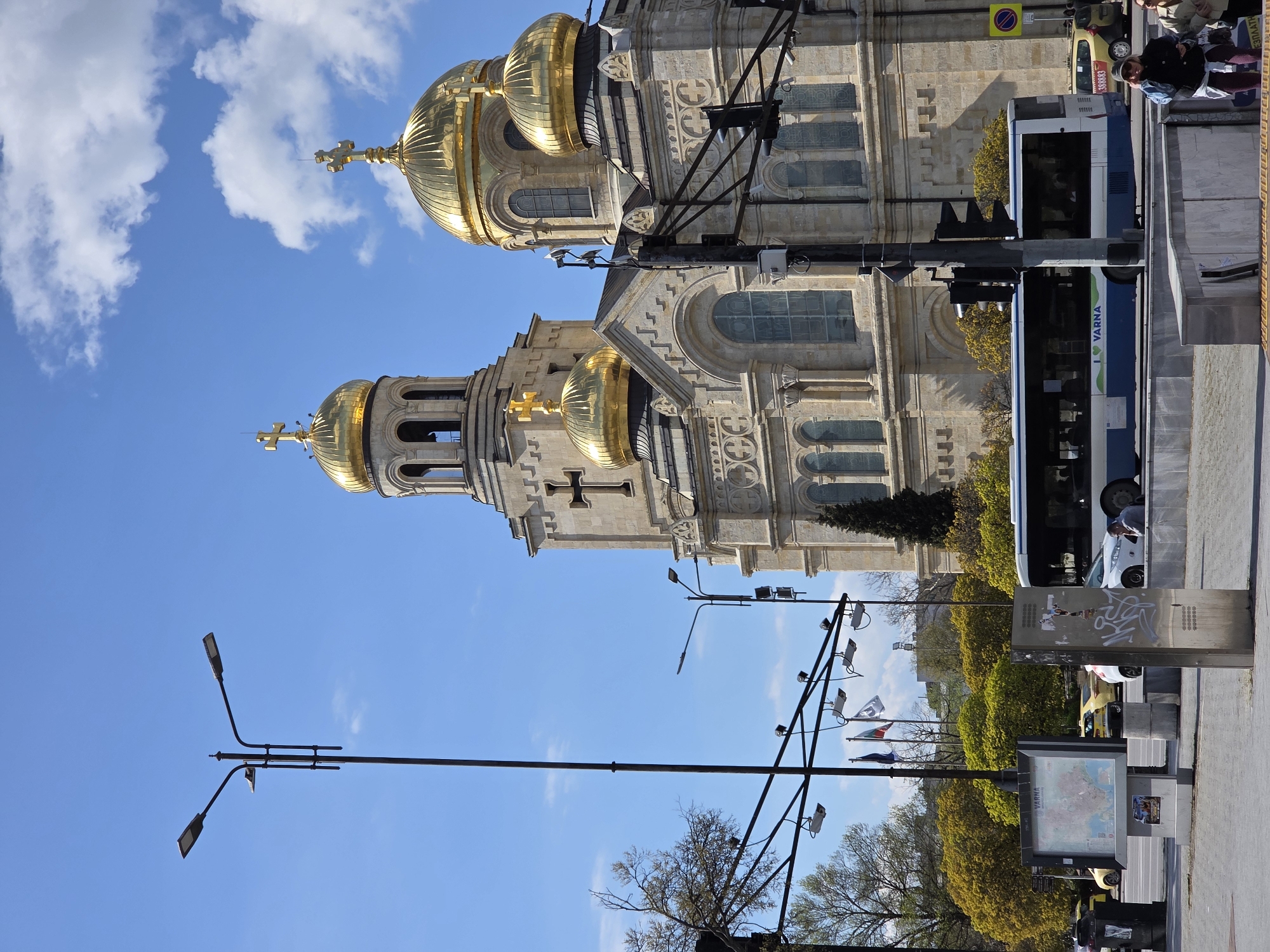 A vibrant daytime street scene in Varna, Bulgaria, showcases the majestic Dormition of the Theotokos Cathedral dominating the left side of the frame. The cathedral is a grand, ornate Eastern Orthodox church, characterized by multiple golden, ribbed domes topped with golden crosses, light stone facade, arched windows, and intricate architectural details including reliefs and a bell tower. The sky above is a clear blue with scattered white clouds.
On the right, a busy city street unfolds. A large public transport bus, primarily blue and white, is prominently featured, displaying "I ❤️ VARNA" on its side, confirming the location. Several people are visible, including one person seated on a bench or low wall near the bus stop, looking towards the camera, and others inside the bus. Behind the bus, a yellow car, possibly a taxi, is partially seen, with "38883" visible on its side.
The street is equipped with typical urban infrastructure, including tall black streetlights, traffic lights, and a robust pole system fitted with multiple security cameras. Further along the pavement, a double-sided information kiosk displays a map with the word "VARNA" clearly visible. Lush green and yellowish-green trees line the street, with some bare branches also present. A yellow circular sign with a red diagonal line, indicating "No Parking" or "No Stopping," is visible above the bus. The scene is illuminated by bright sunlight, suggesting a pleasant, clear day.