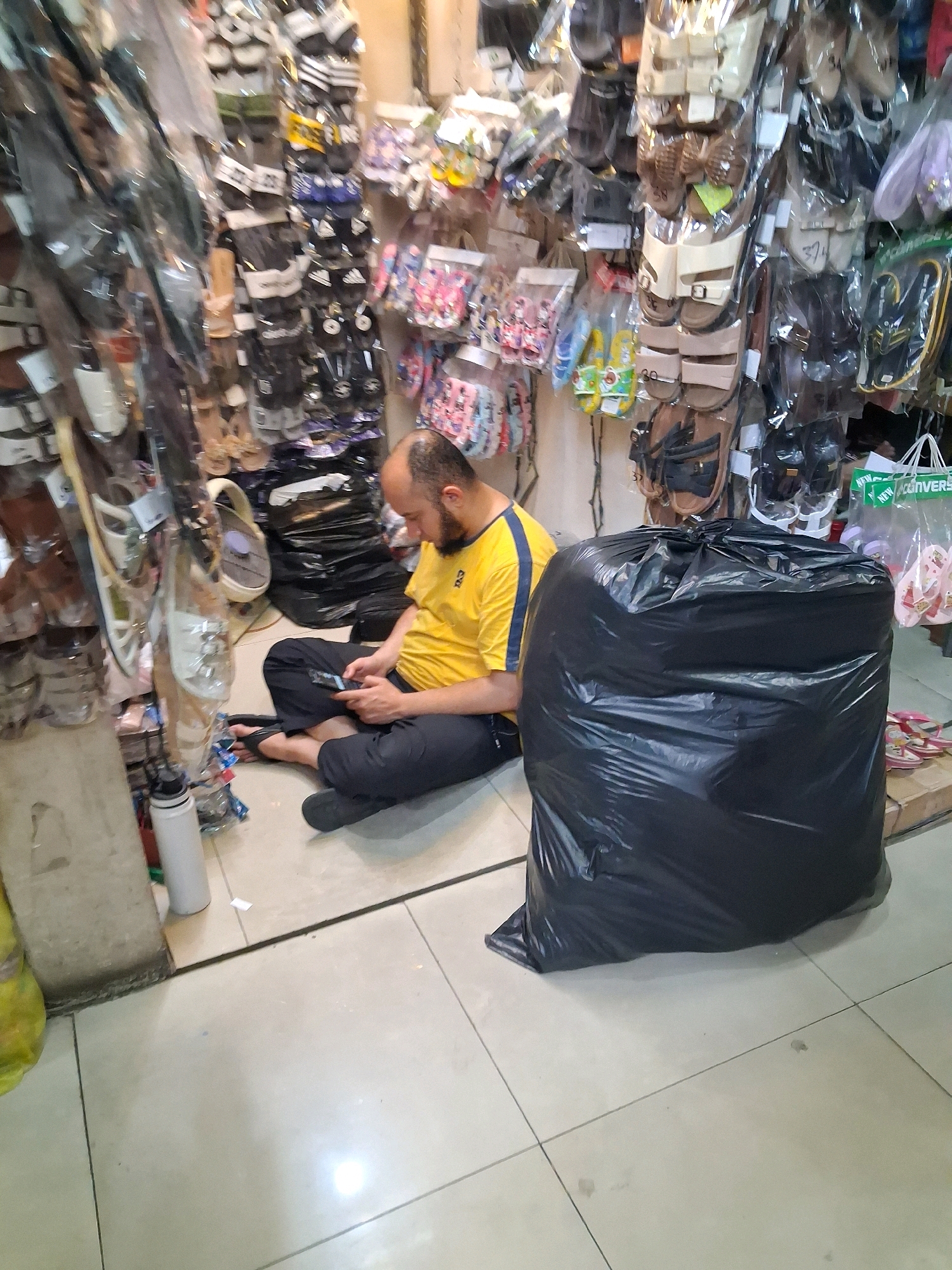 A man is sitting on the floor in a store, using his phone. He is wearing a yellow shirt with blue stripes and black pants. In front of him is a large black trash bag. The store appears to be selling footwear, with many pairs of shoes and sandals displayed on racks and hanging from the ceiling. The floor is tiled.