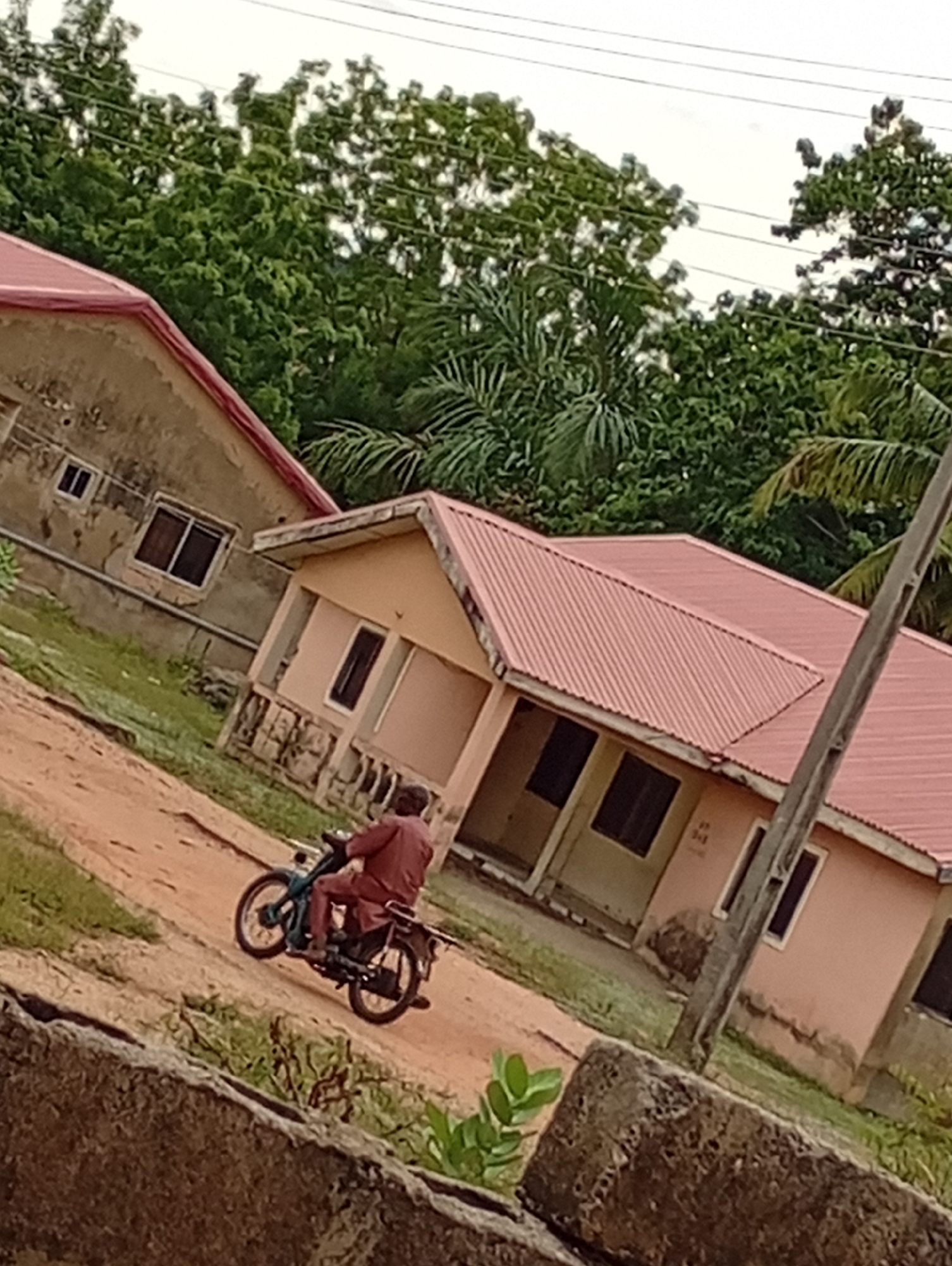 The image depicts a residential area in an unknown location. A person is riding a motorcycle on a dirt road, with buildings visible in the background. The buildings appear to be single-story structures with red roofs. The environment includes trees and vegetation. A concrete structure is visible in the foreground.