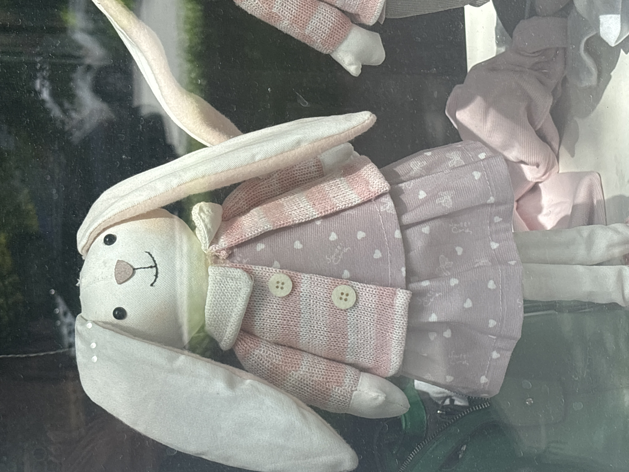 A close-up shot shows a stuffed bunny doll with large, floppy ears, wearing a pink and white striped cardigan over a pink dress adorned with white hearts. The bunny has black button eyes, a stitched smile, and a light green collar. The doll is positioned in front of a window, with reflections of trees and a vehicle visible in the background. The scene appears to be indoors, possibly in a store or home, with a green bag to the left and a pink fabric draped near the doll's feet. The overall impression is that of a display or decorative item. The image does not contain any discernible landmarks or contextual clues that would allow for identification of its location within the city of Novosibirsk, Russia.