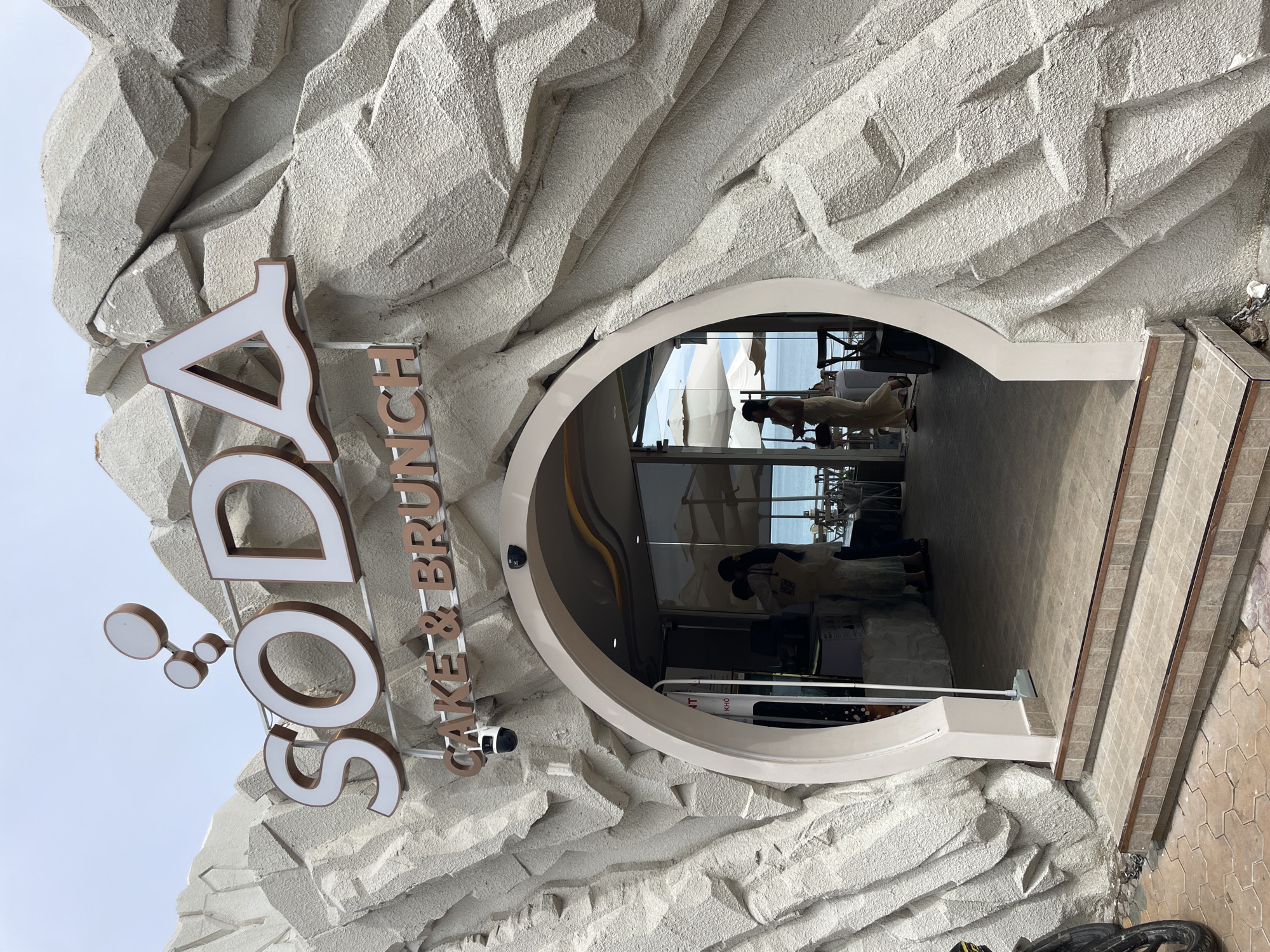 Exterior view of "SODA Cake & Brunch" in Phu Thuy Ward, Vietnam. The restaurant entrance is carved into a textured, off-white facade resembling rock formations. A circular opening serves as the entryway, revealing the interior with patrons inside. A sign bearing the name of the establishment is attached to the facade. Tile steps lead up to the entrance. The weather seems bright and the sky is visible in the background.