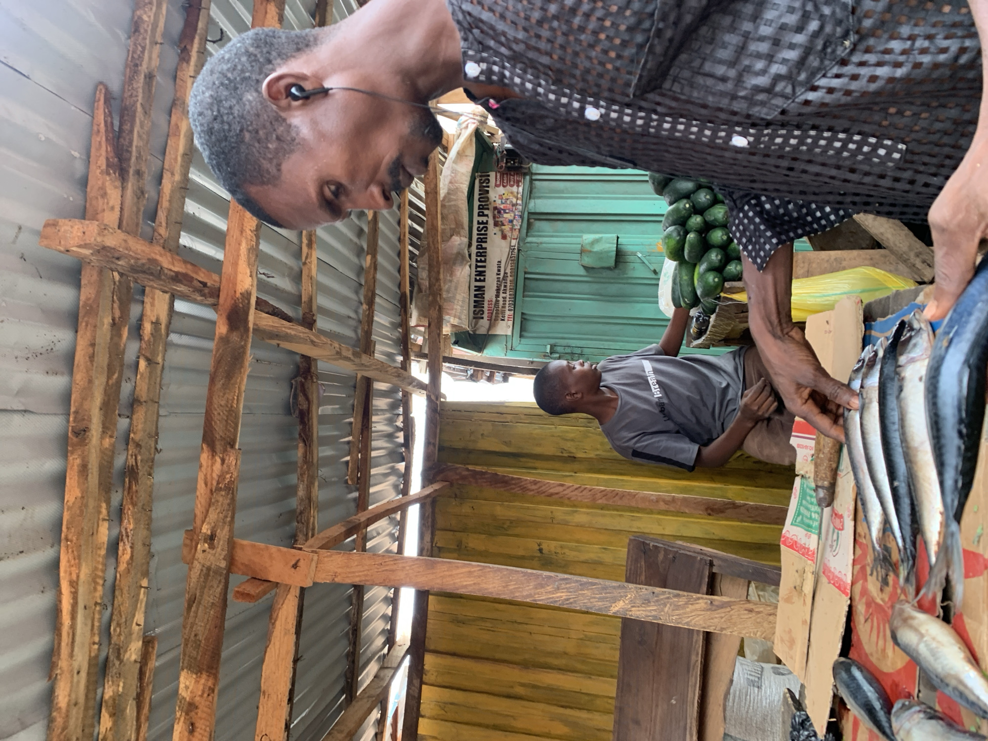This image captures a vibrant market scene in Akwanga, Nigeria, depicting two men and their wares within a rustic stall. In the foreground, a man with short, dark hair and earphones is seen from a slightly elevated angle, wearing a dark checkered shirt. He leans forward, his right hand near a display of several silvery fish, likely mackerel, arranged on a cardboard box for sale.
Behind him, another man, possibly younger, is seated on a low stool or box. He wears a grey t-shirt with the partial text "ban Breath" visible, and looks upwards towards the activities in the foreground.
The stall itself is constructed from rough-hewn wooden planks forming a framework, with corrugated metal sheets serving as walls and possibly roofing. The background features more corrugated metal, painted in shades of yellow and green. To the left, a bunch of green gourds or cucumbers hangs, indicating the sale of fresh produce. A yellow plastic bag is also visible near the produce.
A beige banner prominently displays the business name "ISMAN ENTERPRISE PROVISION," along with an address: "Opposite Makeran Kwata, Keffi Road, Akwanga," and two telephone numbers: "07030878716" and "09013851761." A smaller white sign mentions "CASH" among other less legible text. The bright, natural lighting suggests it is daytime. The overall scene conveys a busy, informal market environment focused on daily commerce.