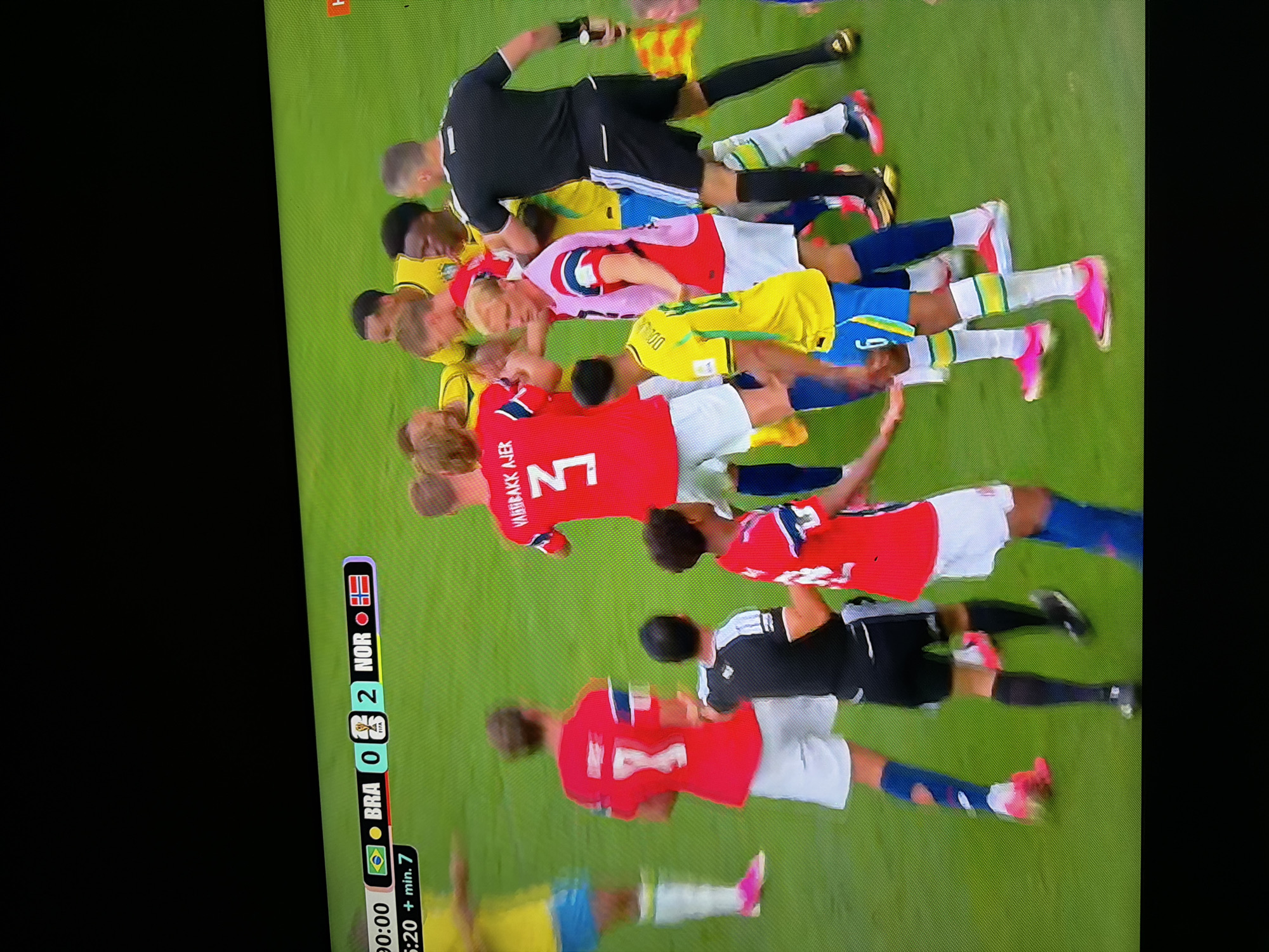 The image depicts a soccer match. The scoreboard at the top left of the image shows "00:00" and the score "BRA 0 2 NOR". The players are wearing red and yellow jerseys. In the foreground, several players are in a huddle, some appearing to be in a confrontation. The environment is a green soccer field. The image does not provide any information about the city of Ognianovo, Bulgaria.