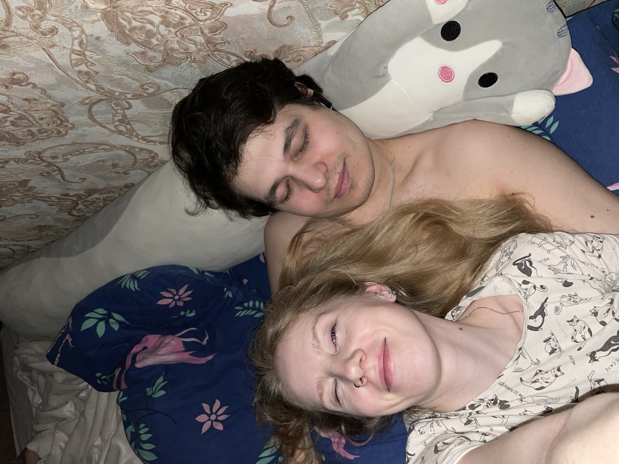 A young adult male and female are lying in bed. The male is asleep with dark hair and a silver chain around his neck. He is shirtless. The female has blonde hair and is wearing a shirt with cat designs. She has a nose ring and multiple ear piercings and is smiling. A stuffed grey and white animal toy is near the male's head. The bedding is blue with a pink flower and flamingo pattern. The location is unknown, but the prompt specified Izhevsk, Russia. Therefore, the location is presumed to be Izhevsk, Russia.