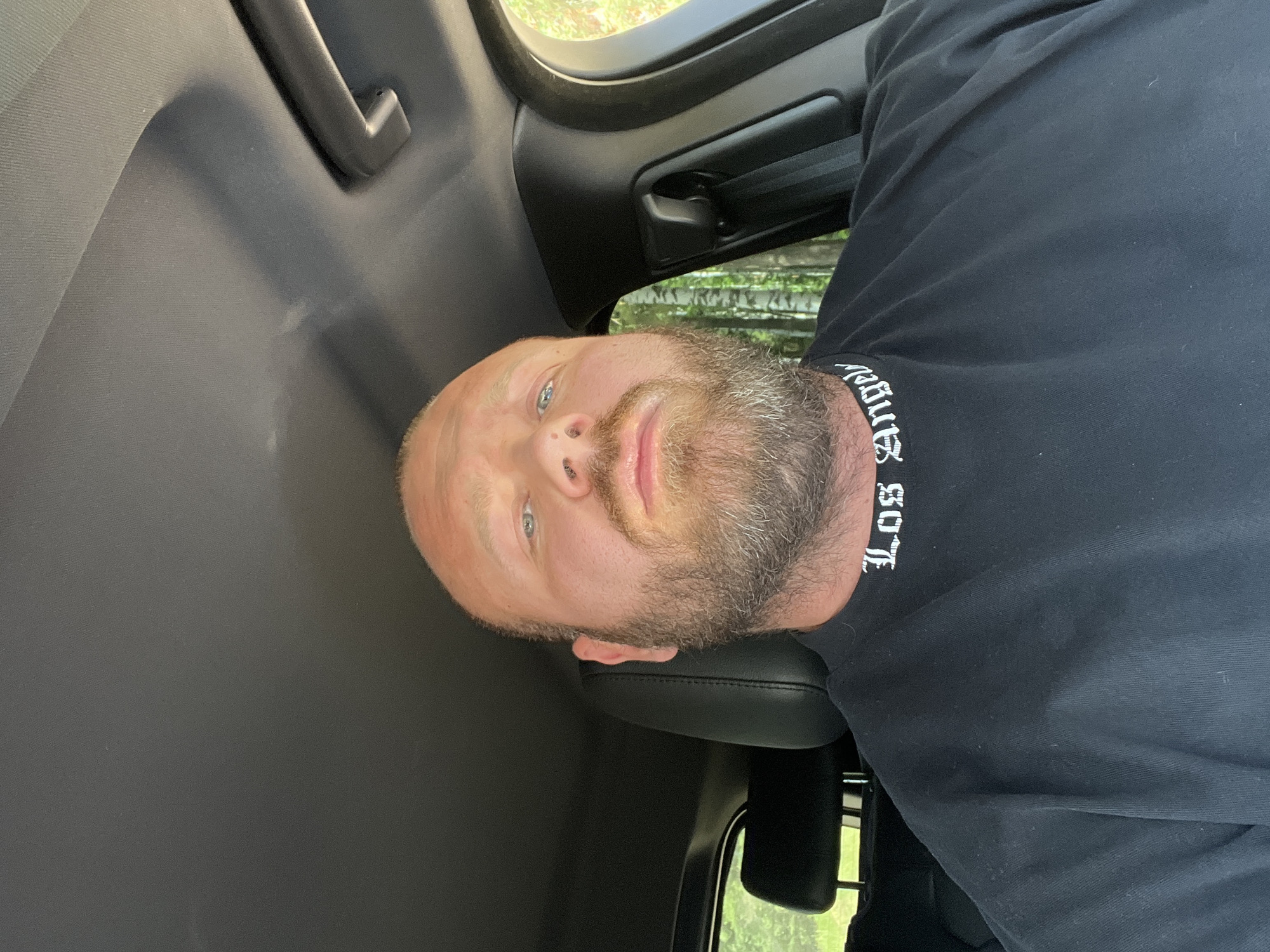 The image shows a close-up selfie of a bald, light-skinned man with a beard, seated in the driver's seat of a car. He is wearing a black t-shirt with "Los Angeles" written in white gothic font around the collar. The interior of the car, including the dashboard and headliner, is visible. Outside the car window, a forest with birch trees can be seen, suggesting a rural or natural setting. The image does not provide any specific information about the location in the city of Noviy, Russia.