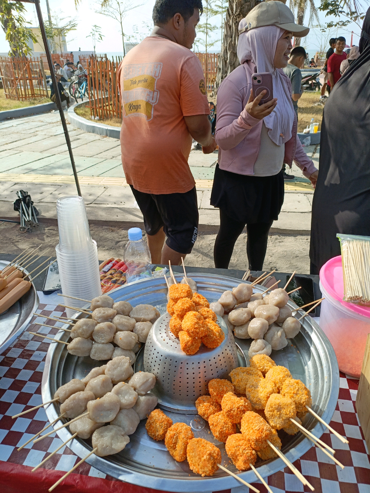 In the city of Bulukumba, Indonesia, a street vendor displays various grilled food items on a table covered with a red and white checkered tablecloth. The vendor's offerings include several skewers of light-colored, round meatballs and skewers of orange, breaded, nugget-like food items. A large metal sieve containing more of the orange food items is positioned behind the meatballs. To the left of the food display are skewers of sausages and a stack of clear plastic cups. A blue plastic water bottle is also visible. In the background, people are seen walking and standing, some in casual attire and others in headscarves, suggesting a public outdoor area. The lighting indicates it is daytime, and palm trees and some fencing are present in the background, implying a coastal or park-like setting.