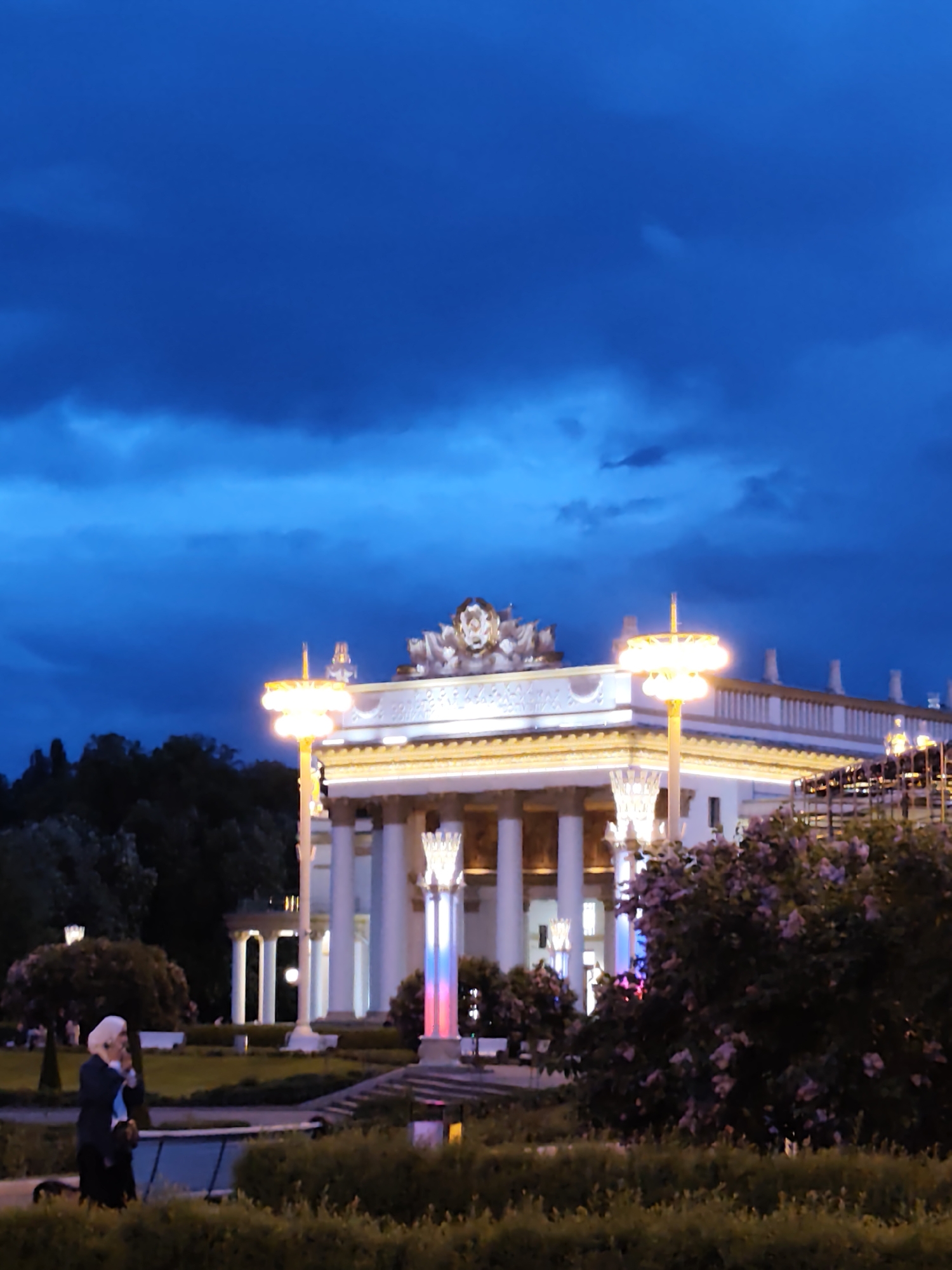 The image shows a building in Moscow, Russia, at night. The building is a white structure with columns and ornate details, illuminated by lights. In front of the building is a garden area with bushes and flowers. A woman wearing a headscarf and a dark coat is walking along a path in the foreground. The sky is a dark blue color. The scene evokes a sense of peace and tranquility.